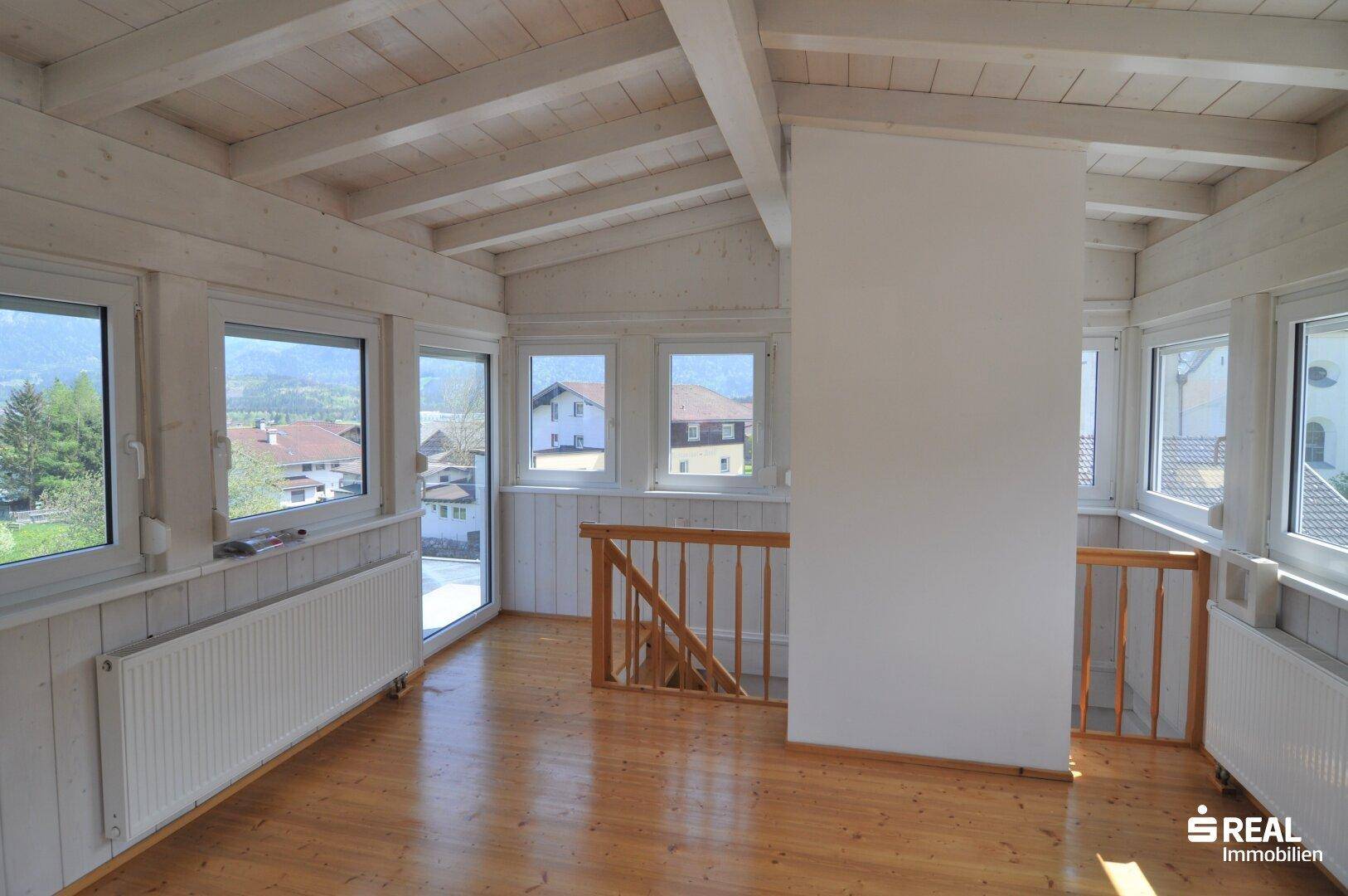 Heller Dachbodenraum mit sichtbaren Holzbalken, vielen Fenstern und Holzfußboden.