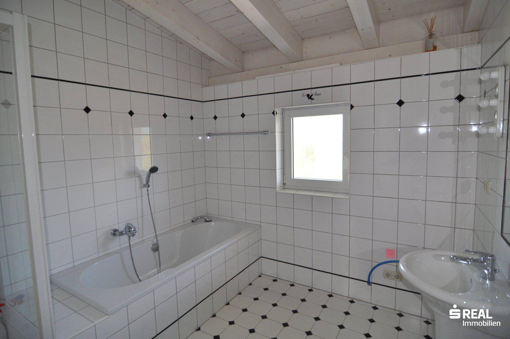 Badezimmer mit Badewanne, Waschbecken und weißen Fliesen mit schwarzem Dekor.