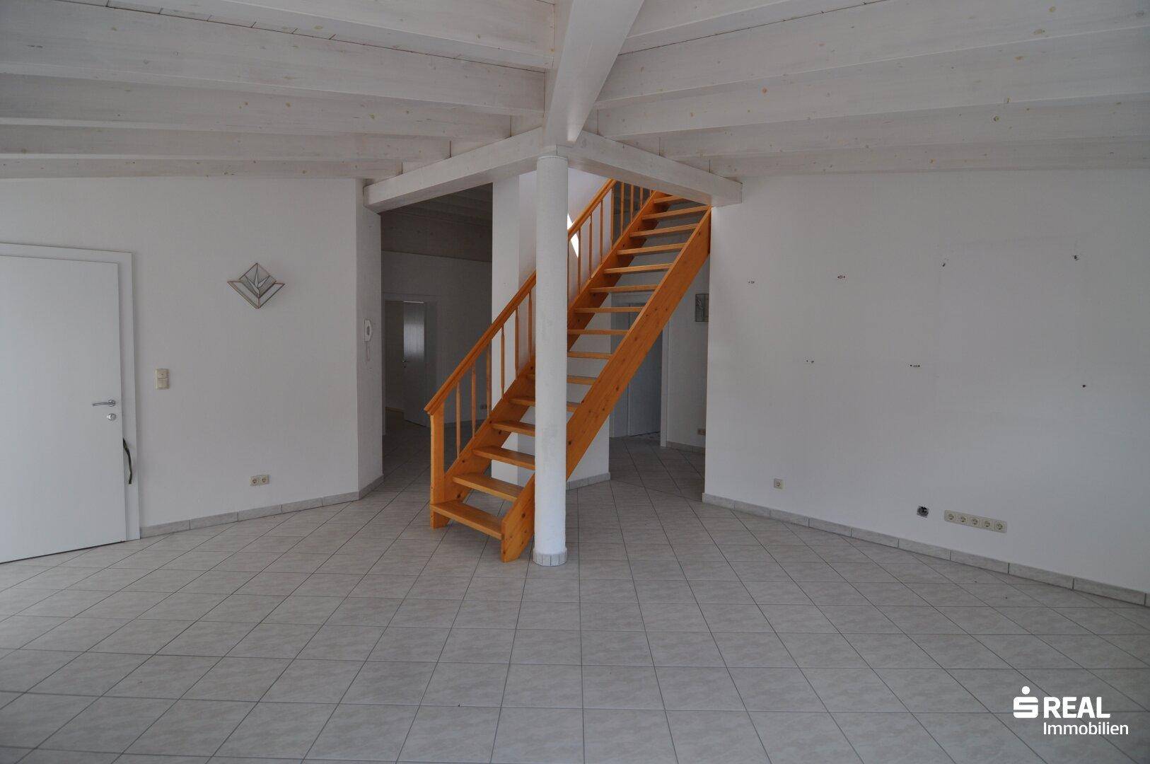 Geräumiger Flur mit Holztreppe, Fliesenboden und weiß gestrichenen Wänden.