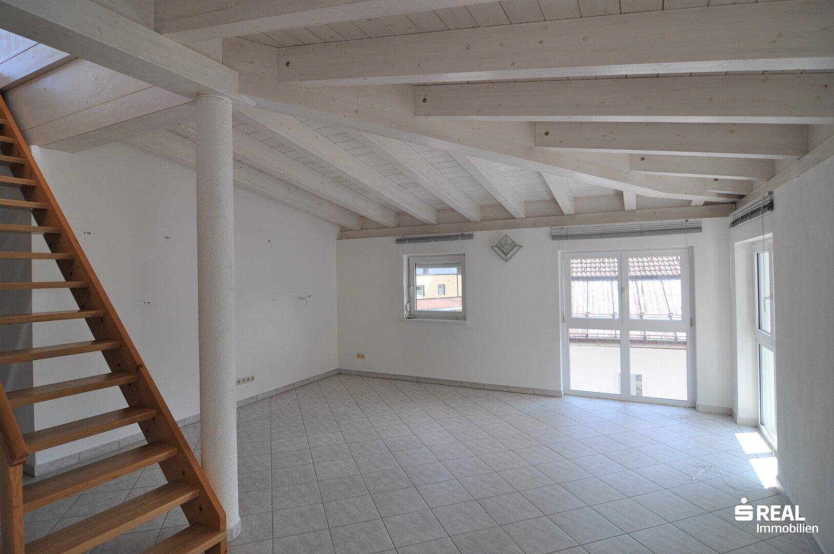 Heller Wohnbereich mit Fliesenboden, Holztreppe und Zugang zu einem Balkon.