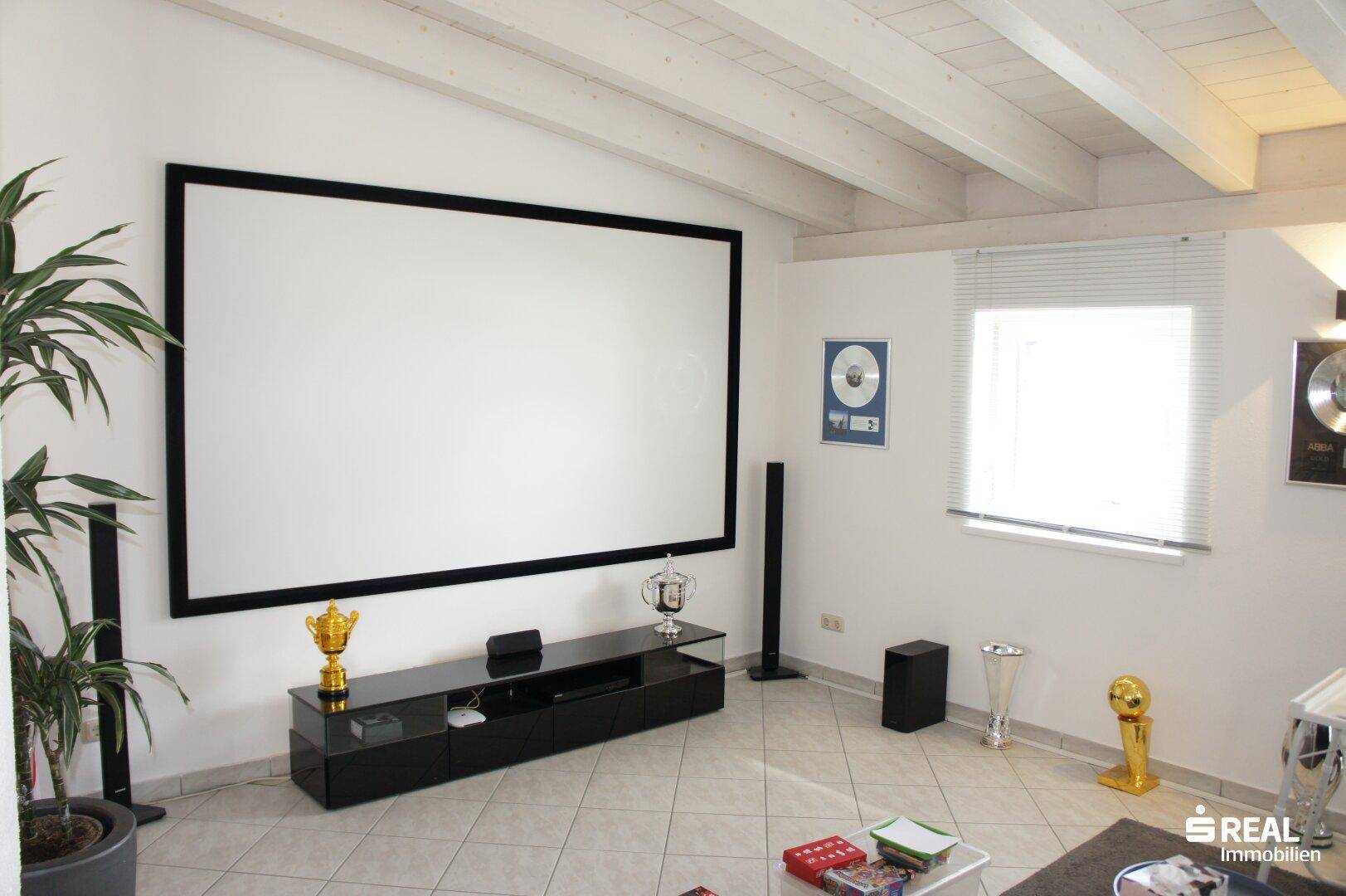 Wohnzimmer mit großer Leinwand, Fliesenboden und sichtbaren Holzbalken an der Decke.