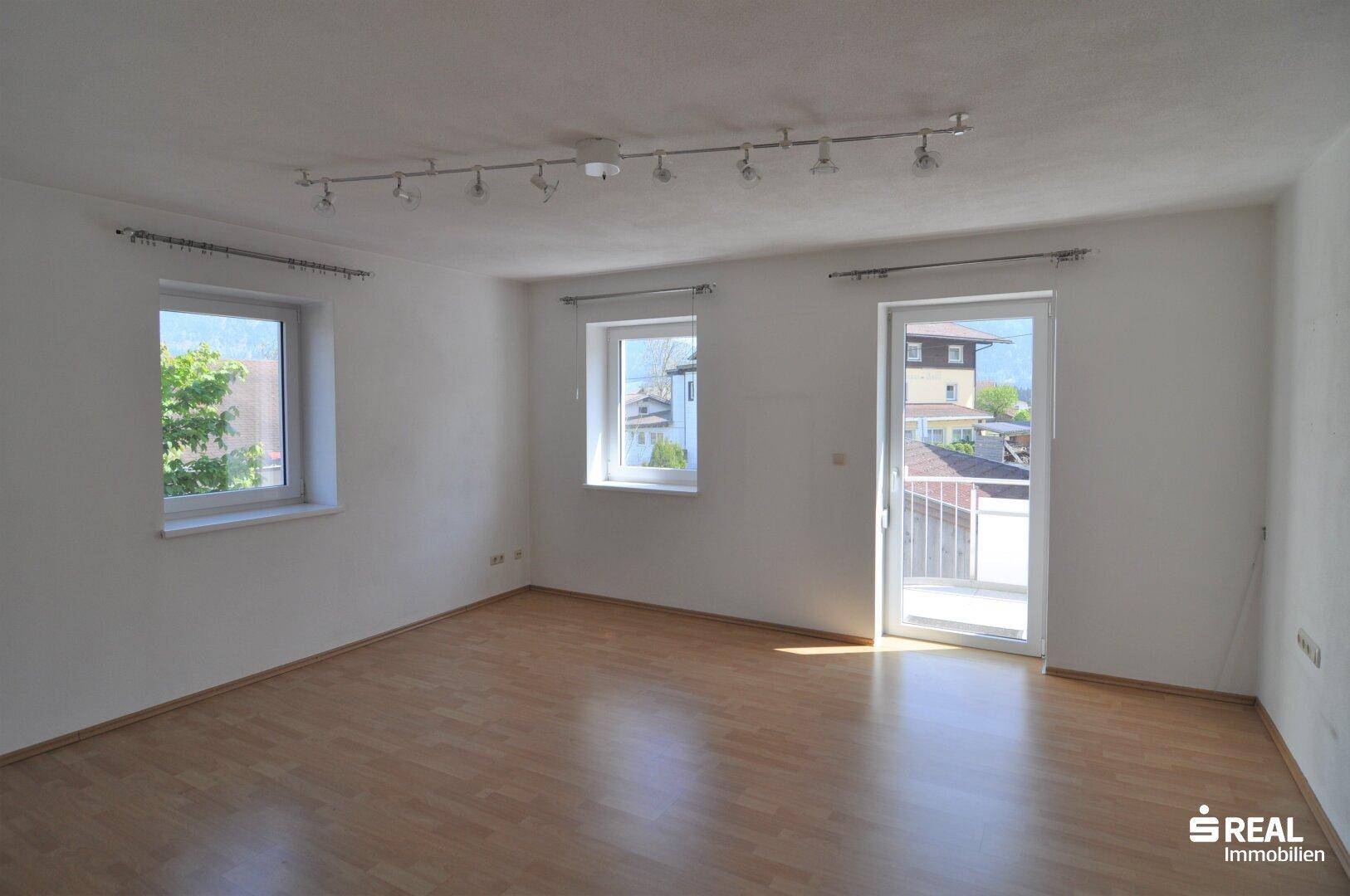 Heller Wohnraum mit Laminatboden, mehreren Fenstern und Zugang zu einem französischen Balkon.