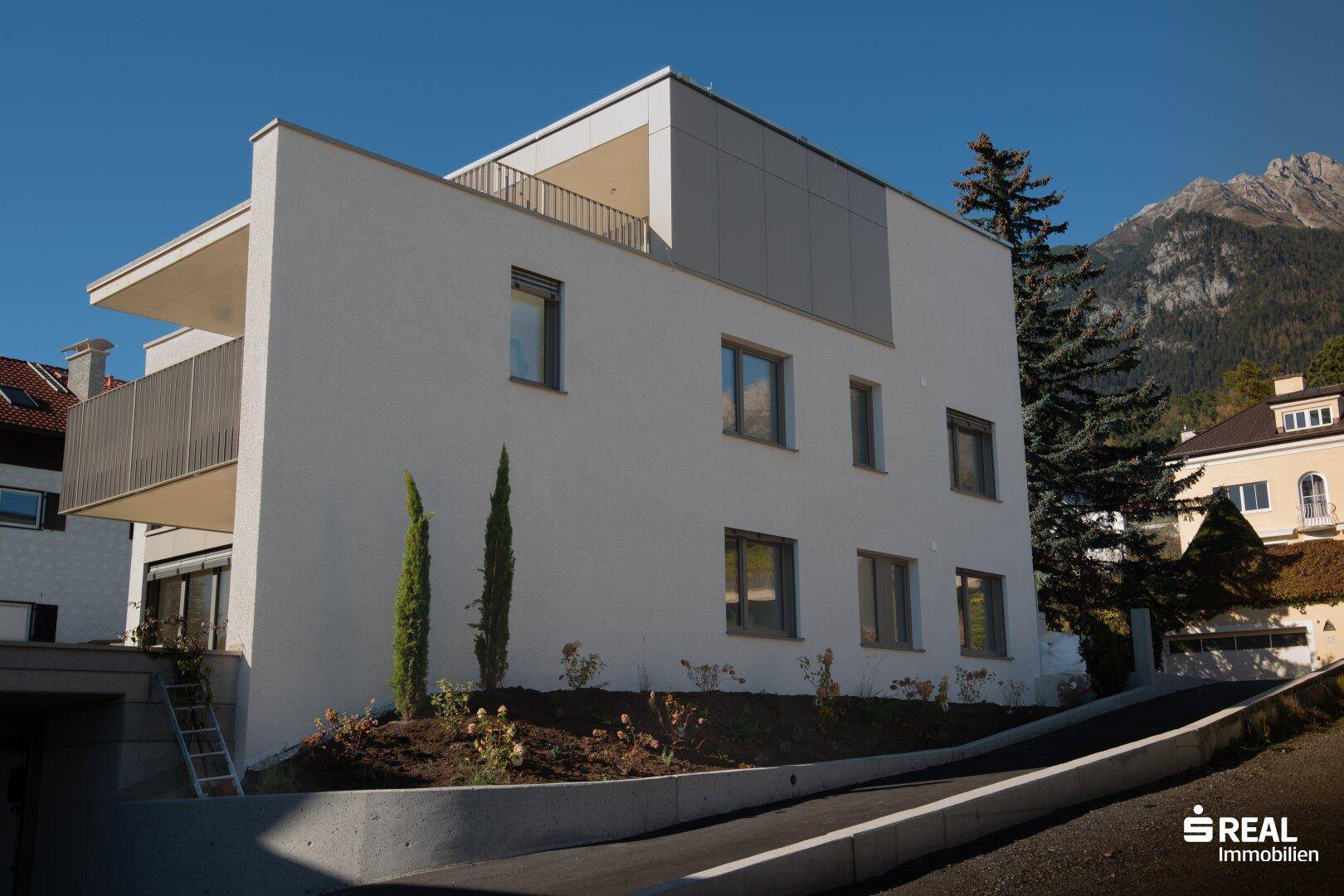 Modernes Gebäude mit weißer Fassade, Zufahrt und Blick auf die umliegenden Berge.