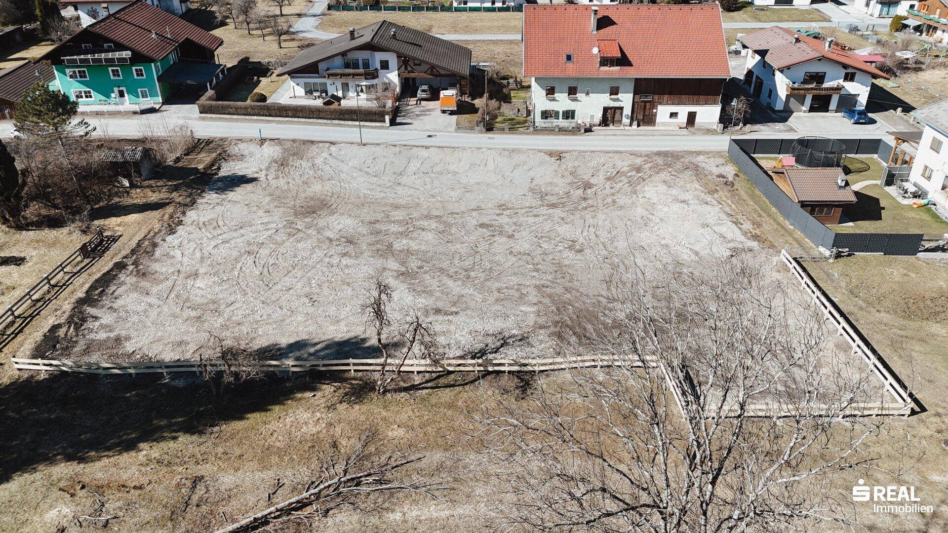 Weitläufige Luftaufnahme eines leeren Baugrundstücks, das von einer Holzumzäunung und weiteren Wohnhäusern begrenzt wird.