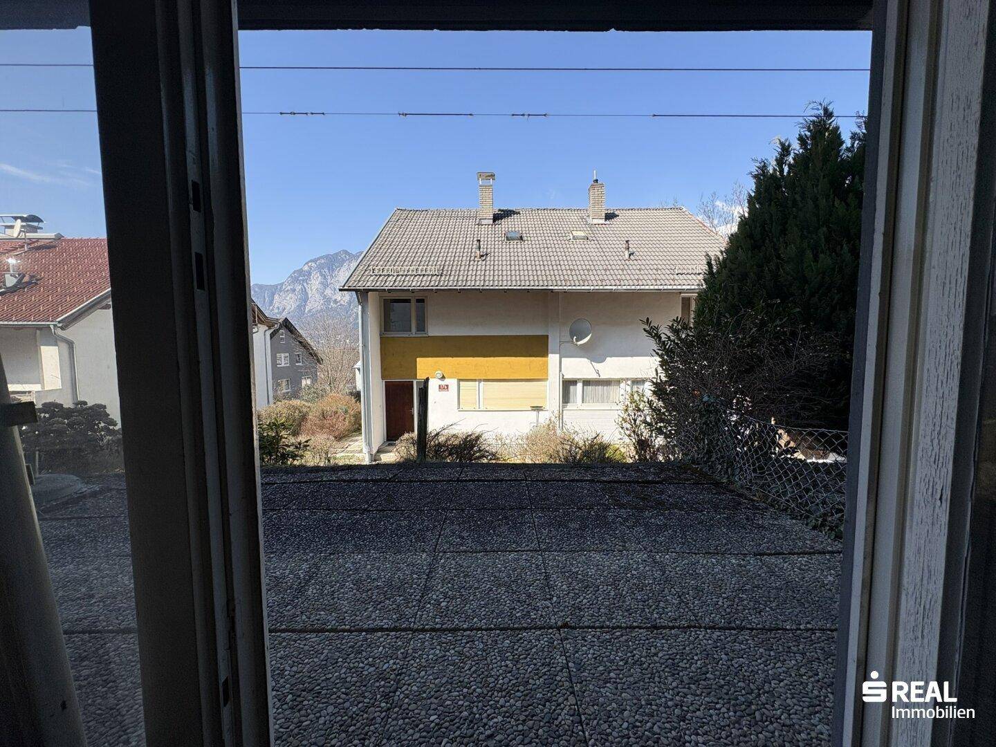 Blick von innen auf eine gepflasterte Terrasse und das gegenüberliegende Haus mit Bergblick.