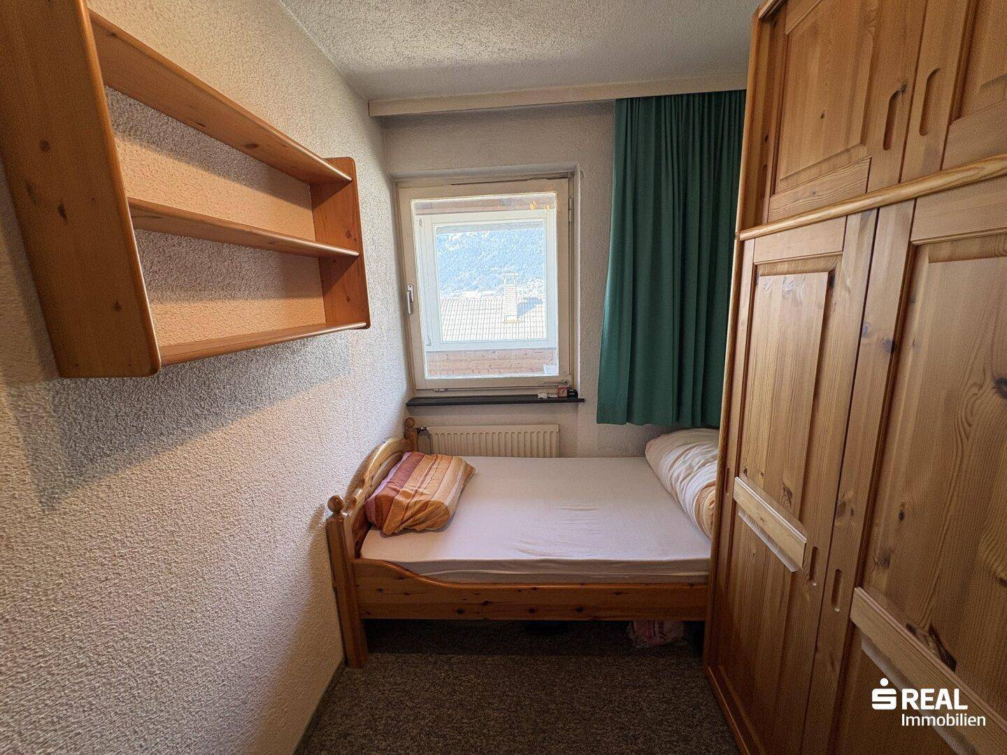 Kleines Schlafzimmer mit Einzelbett, Holzschrank und Fenster mit Blick nach draußen.