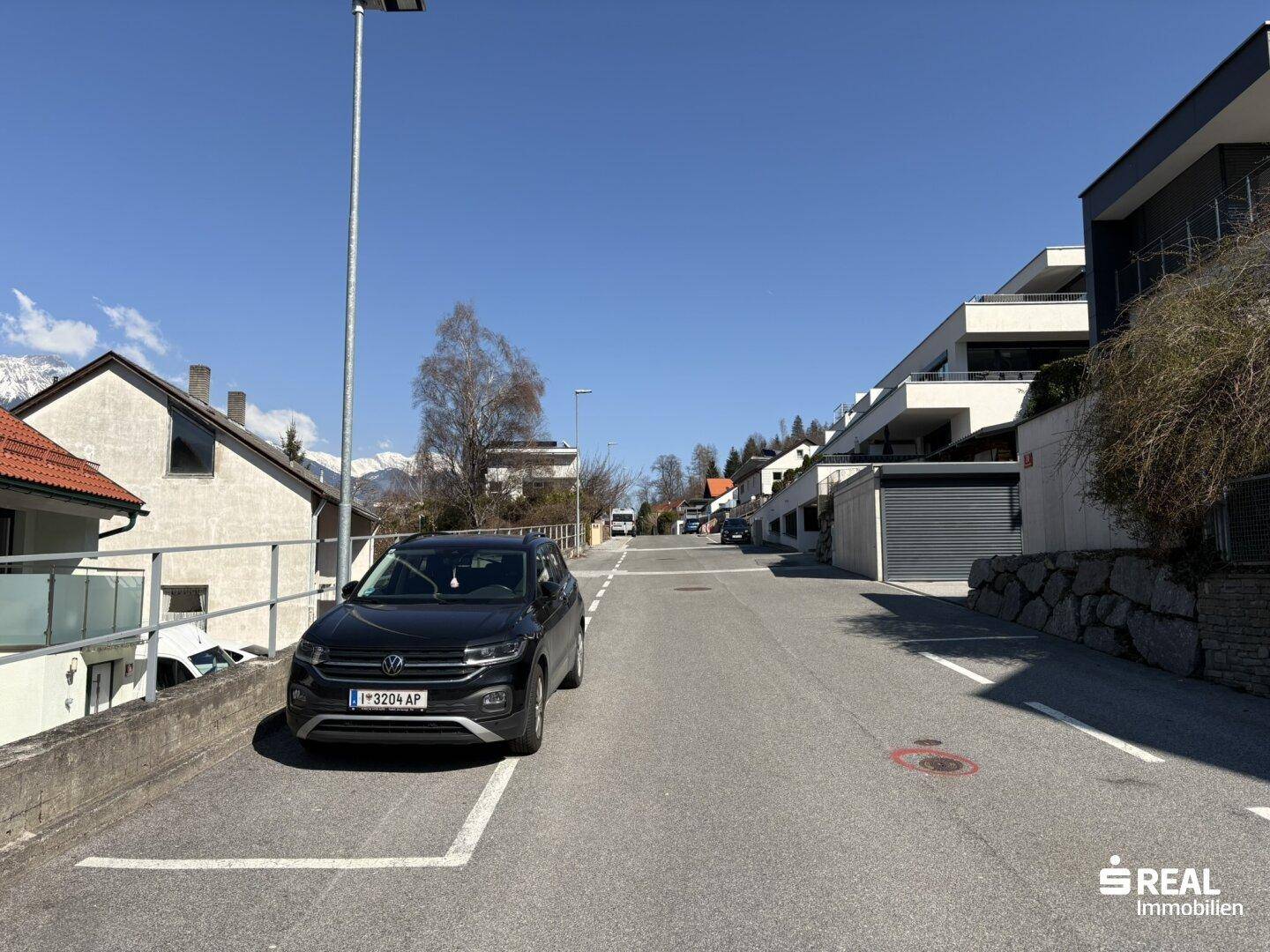 Ruhige Wohnstraße mit geparkten Autos und modernen Gebäuden im Hintergrund, umgeben von Bergen.
