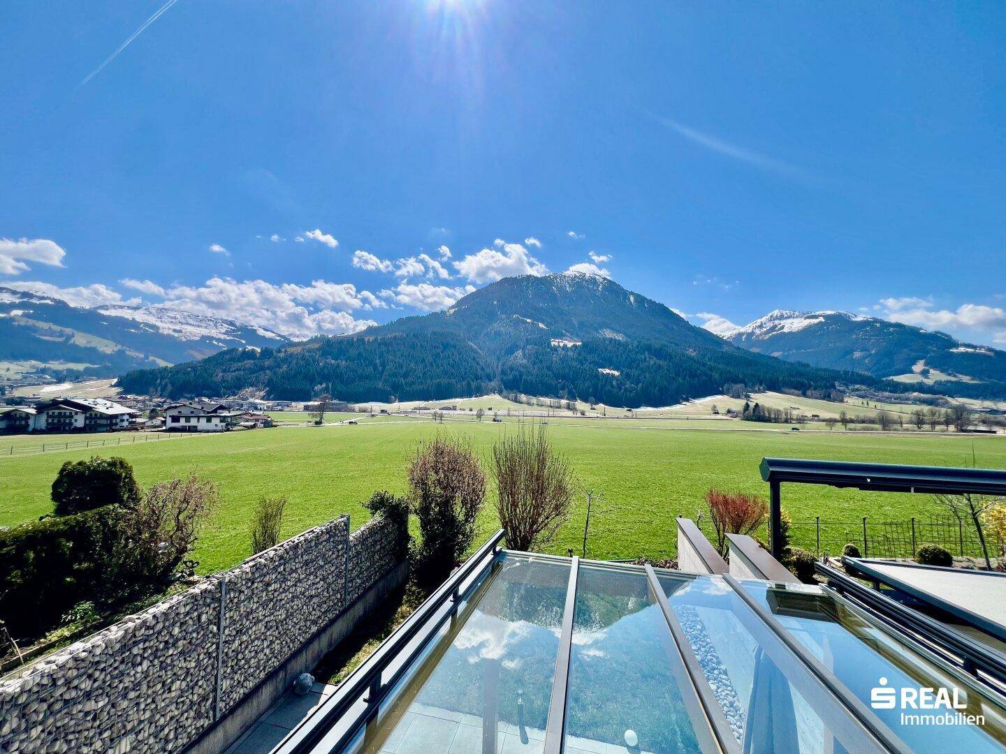 Weitläufiger Panoramablick über grüne Wiesen und schneebedeckte Berge unter blauem Himmel.