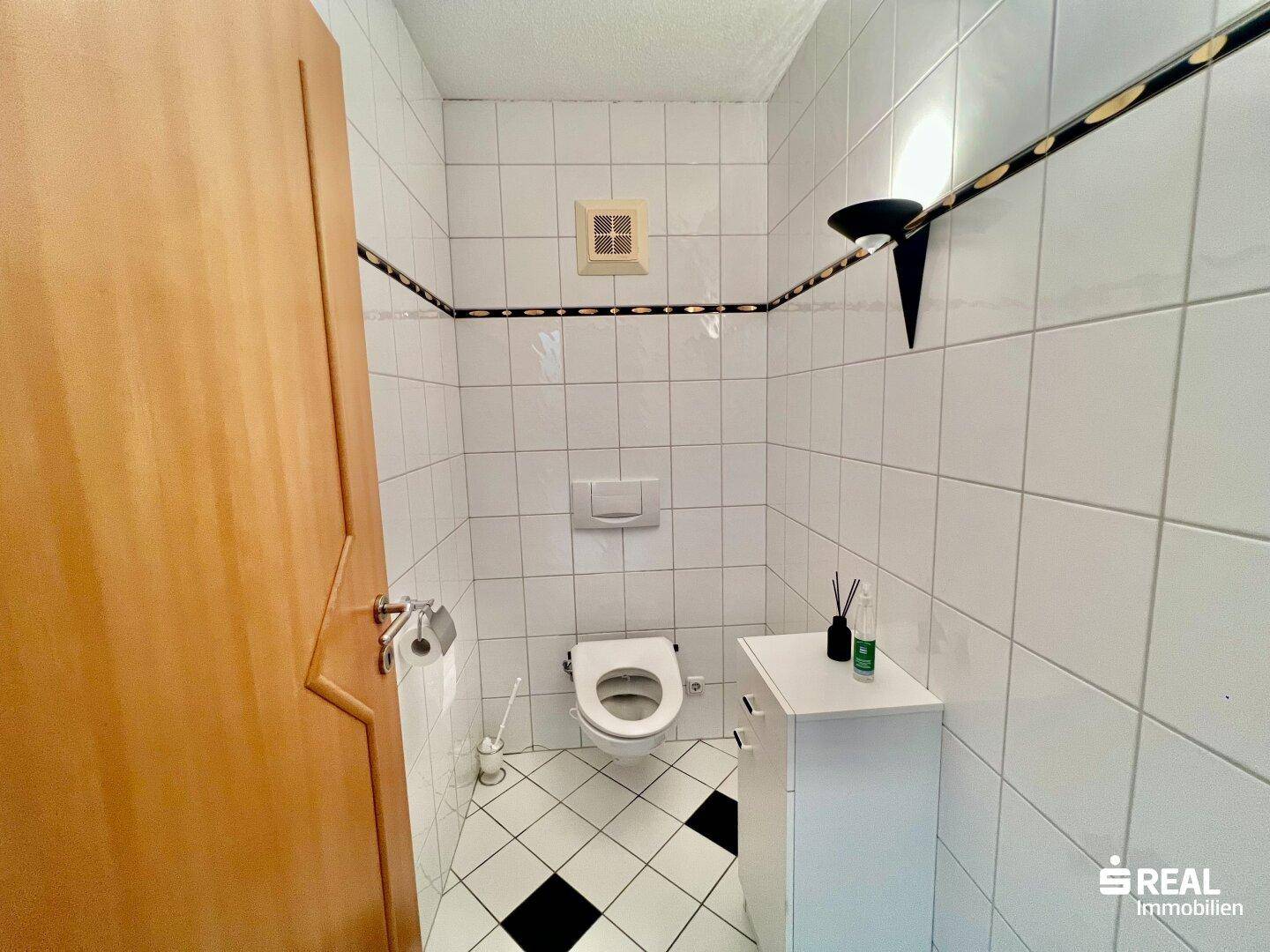 Kleines Gäste-WC mit Toilette und Waschbecken, gefliest in Weiß mit schwarzen Akzenten.