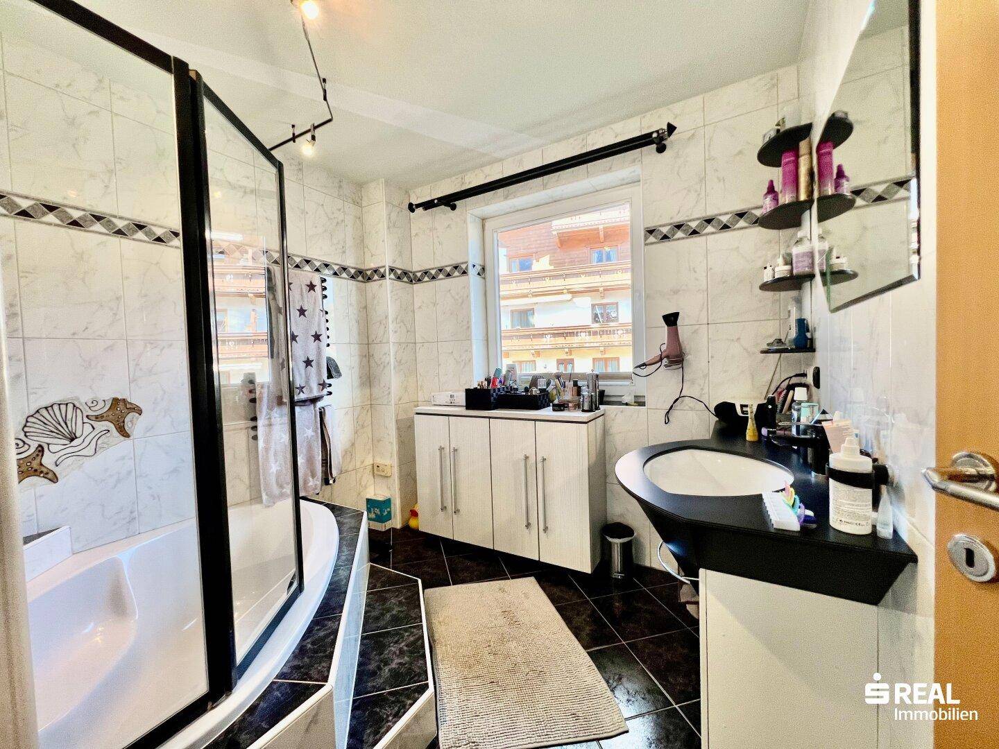 Badezimmer mit Eckbadewanne, Dusche, Waschbecken und Fenster, gefliest in Schwarz-Weiß.