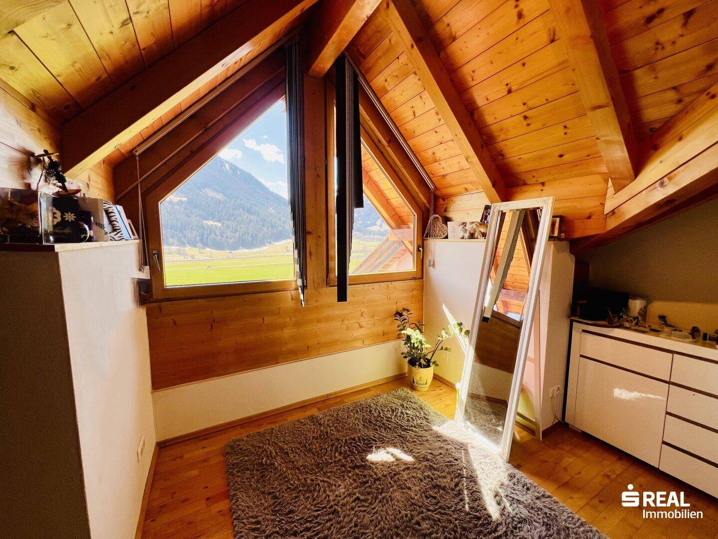 Gemütliches Dachzimmer mit Holzbalken, großen Fenstern und weitem Blick auf die Berge.