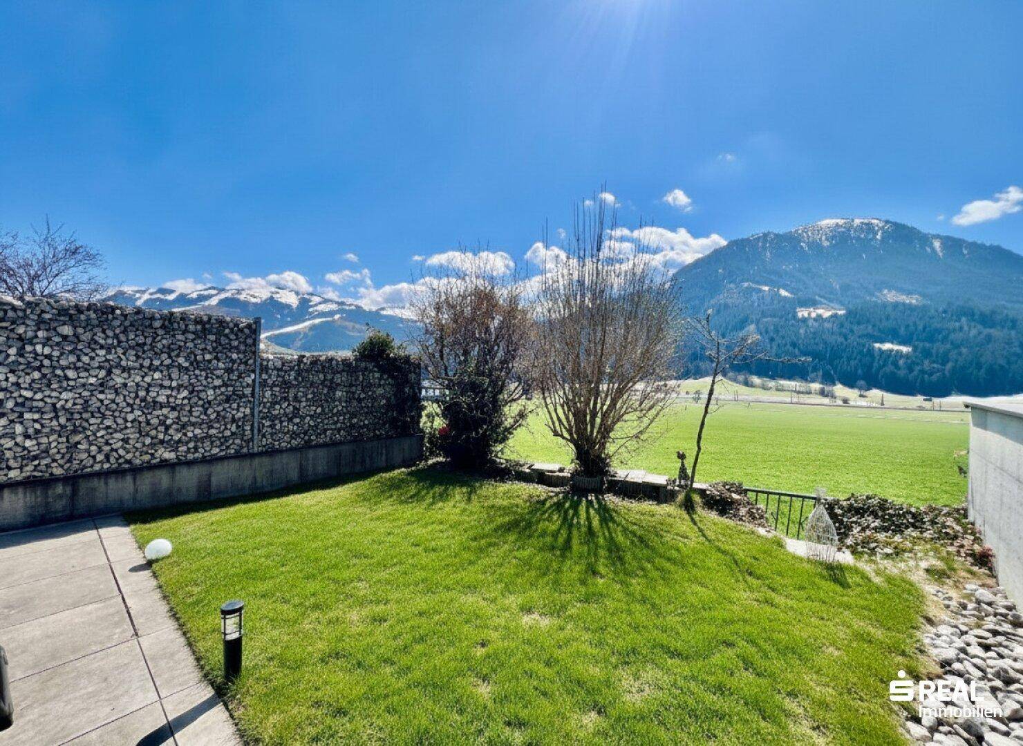 Gepflegter Garten mit Rasenfläche, Gabionenwand und weitem Blick auf die umliegenden Berge.