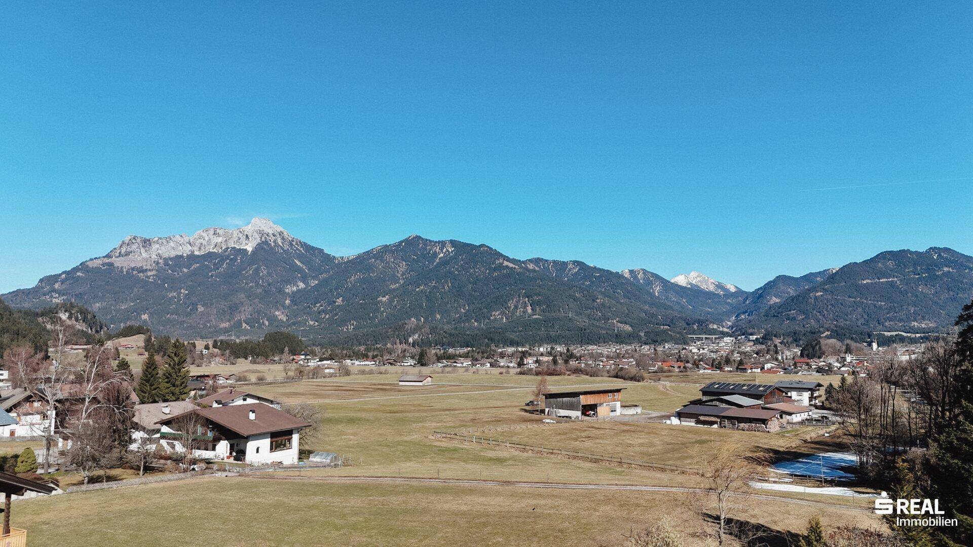 Weitläufiger Blick über das Tal mit Wiesen, Häusern und der beeindruckenden Bergkulisse der Alpen.