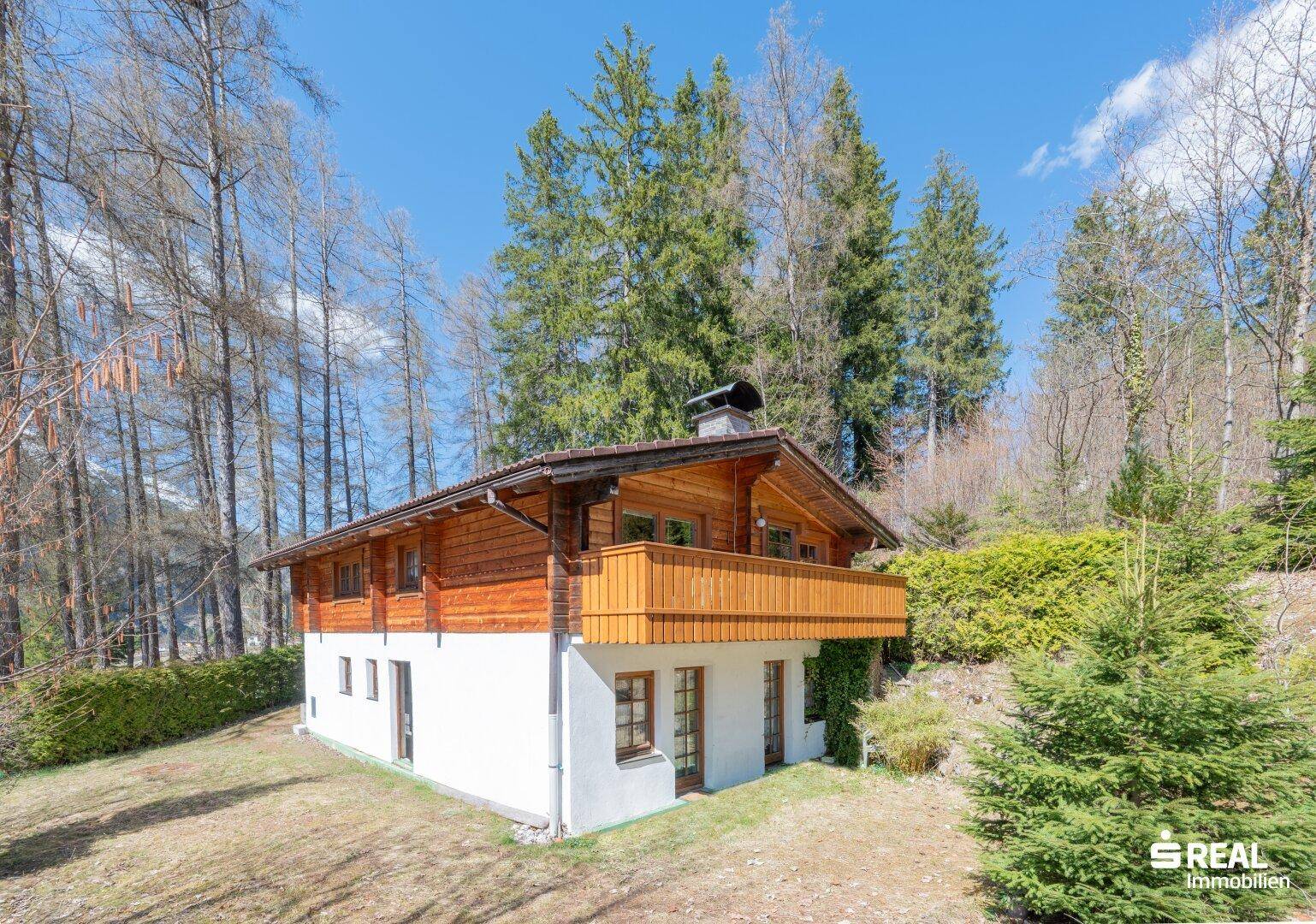 Seitenansicht des Holzhauses mit weißem Sockel und Holzbalkon, umgeben von Bäumen und Natur.
