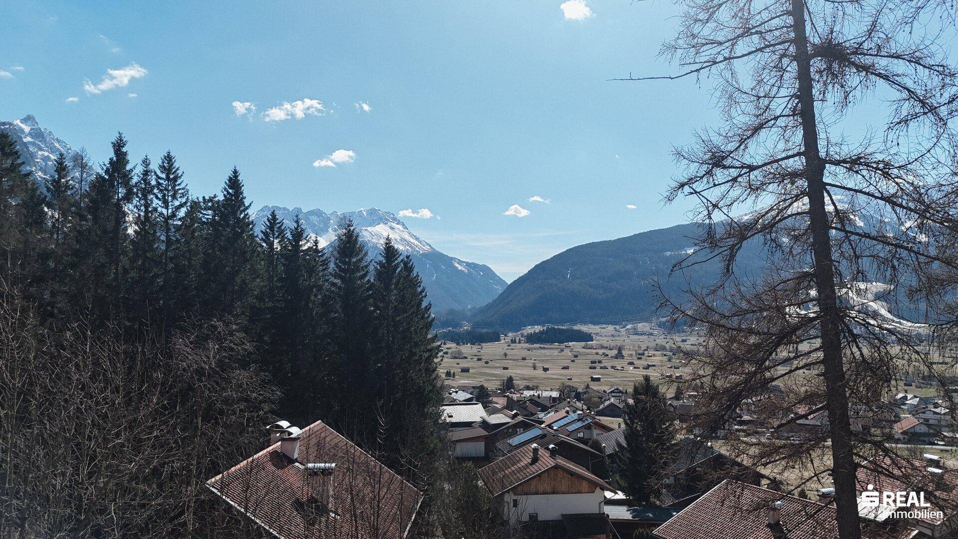 Weitläufiger Blick über ein Tal mit Siedlung und schneebedeckten Bergen am Horizont.