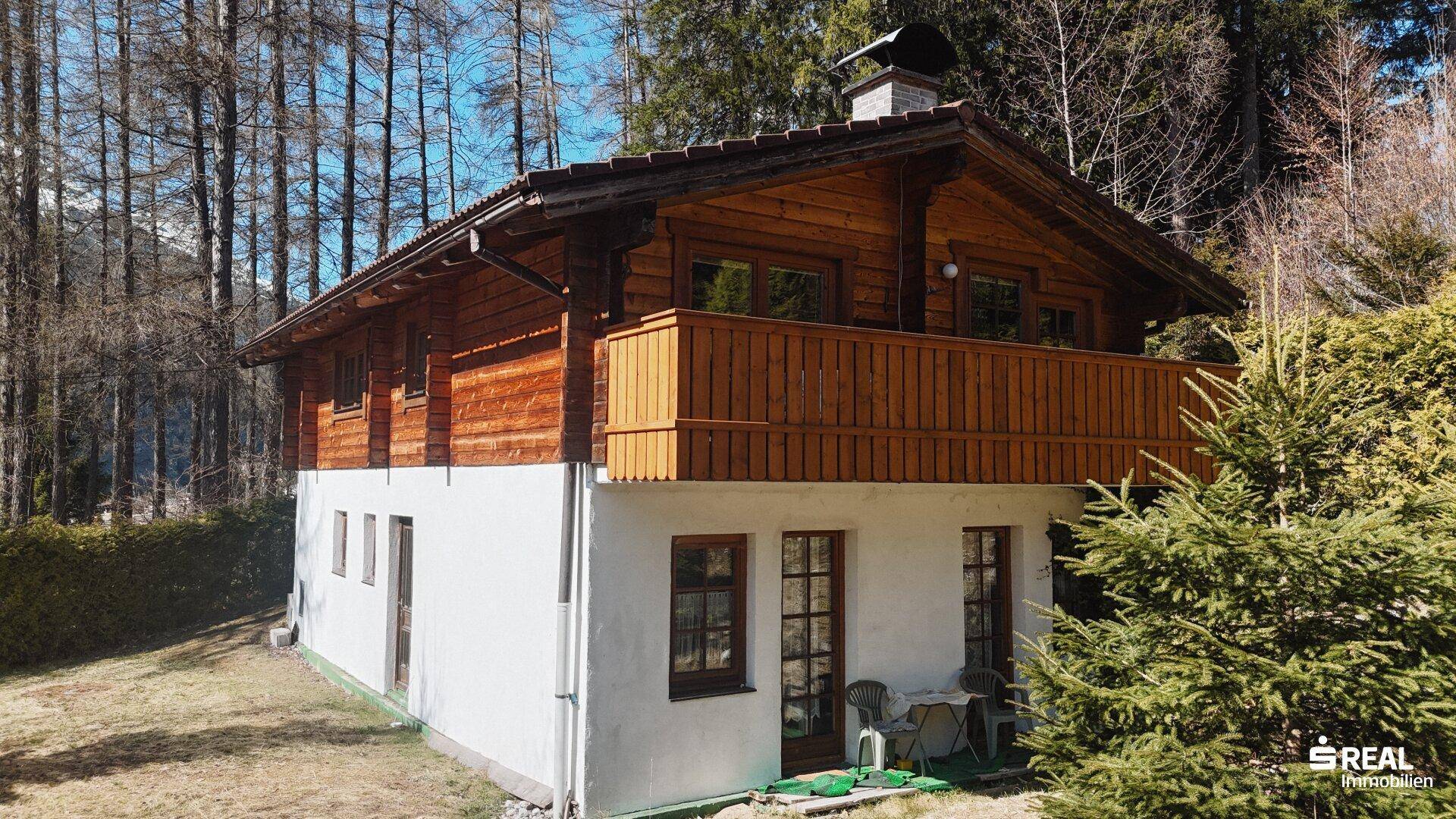Ansicht des Holzhauses mit weißem Sockel und Holzbalkon, umgeben von Bäumen und viel Grün.