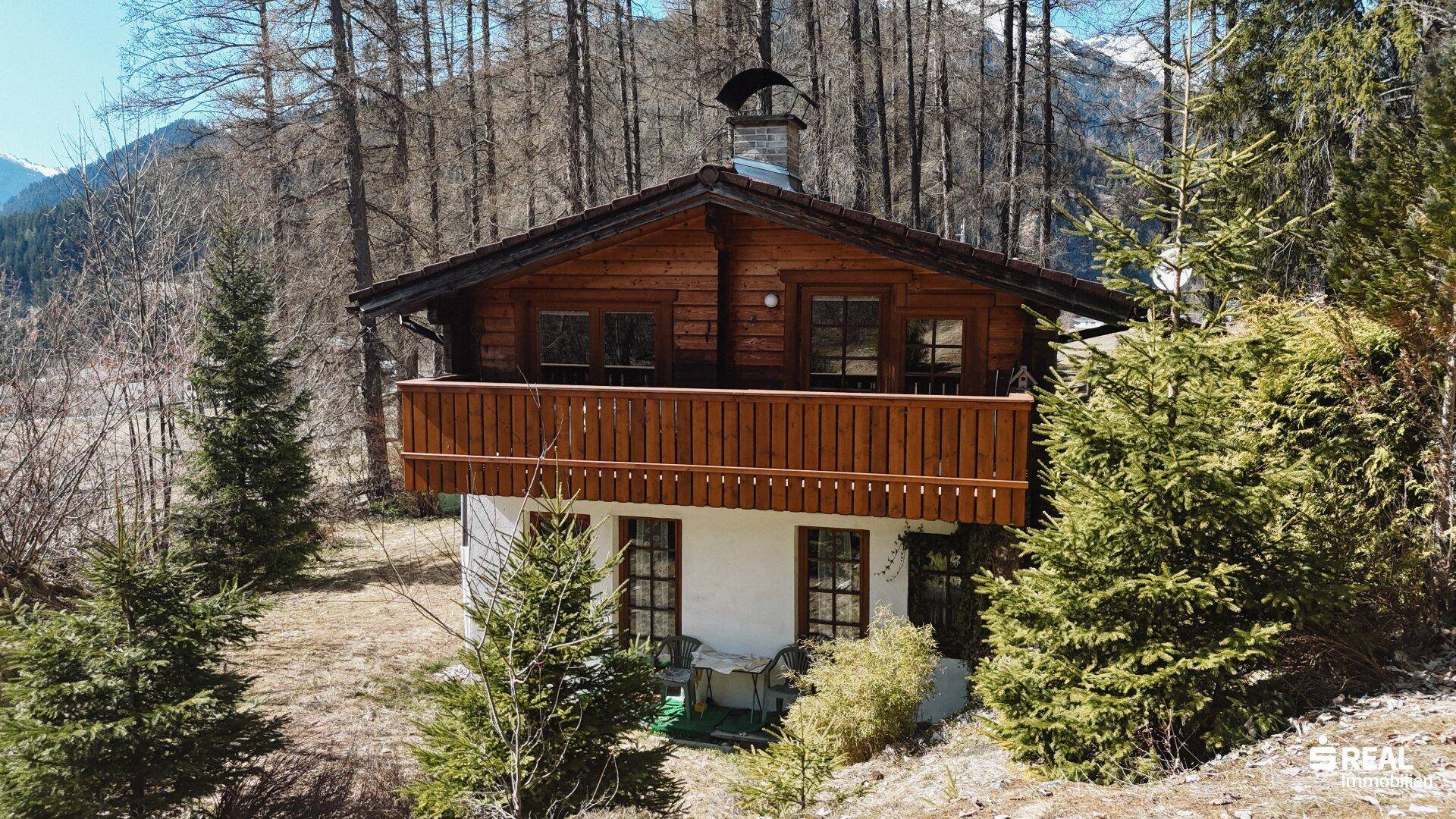 Frontansicht des Holzhauses mit Balkon und Fenstern, umgeben von Nadelbäumen und Natur.