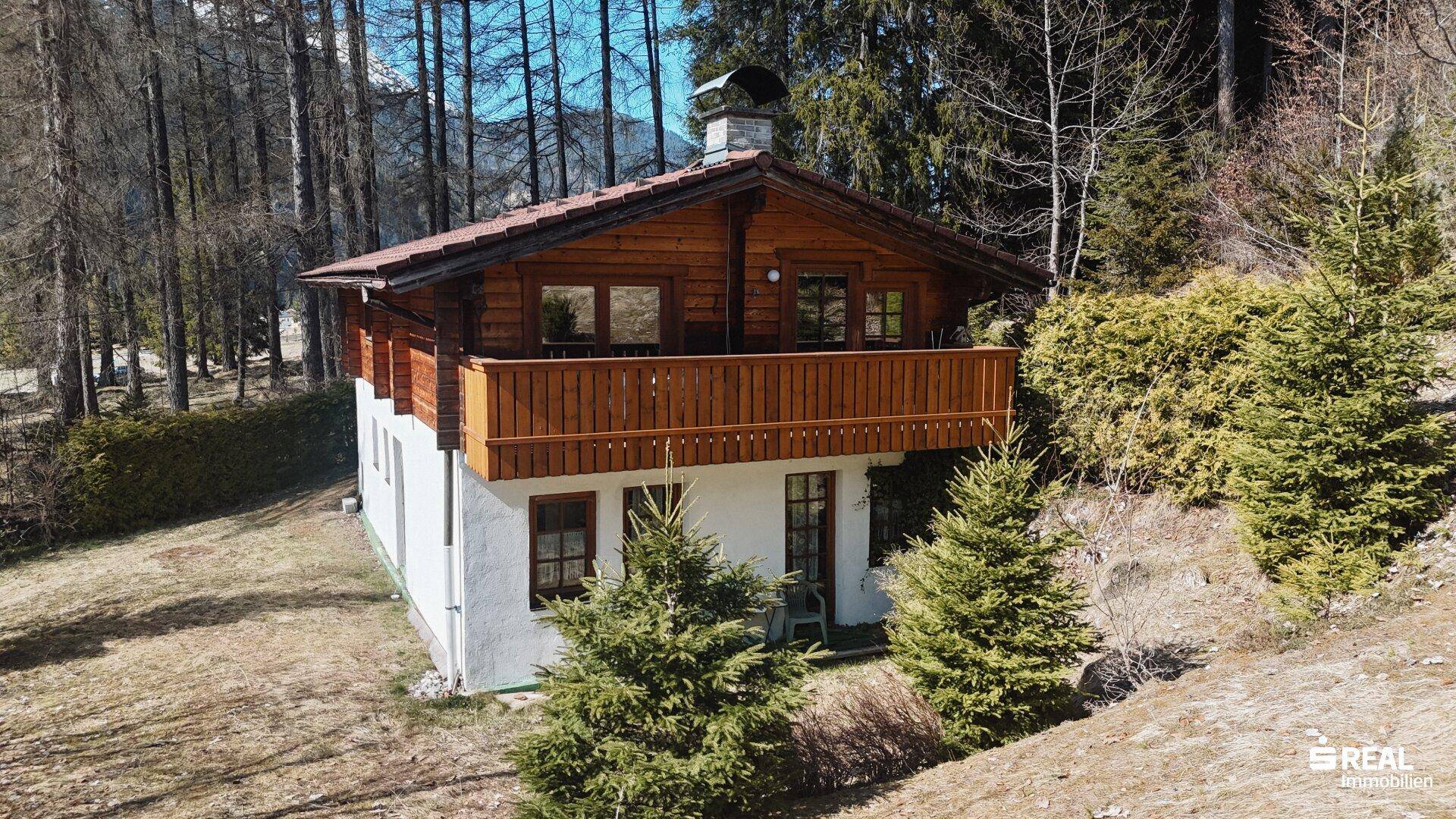 Holzhaus mit Balkon und weißem Sockel, harmonisch in die alpine Landschaft integriert.