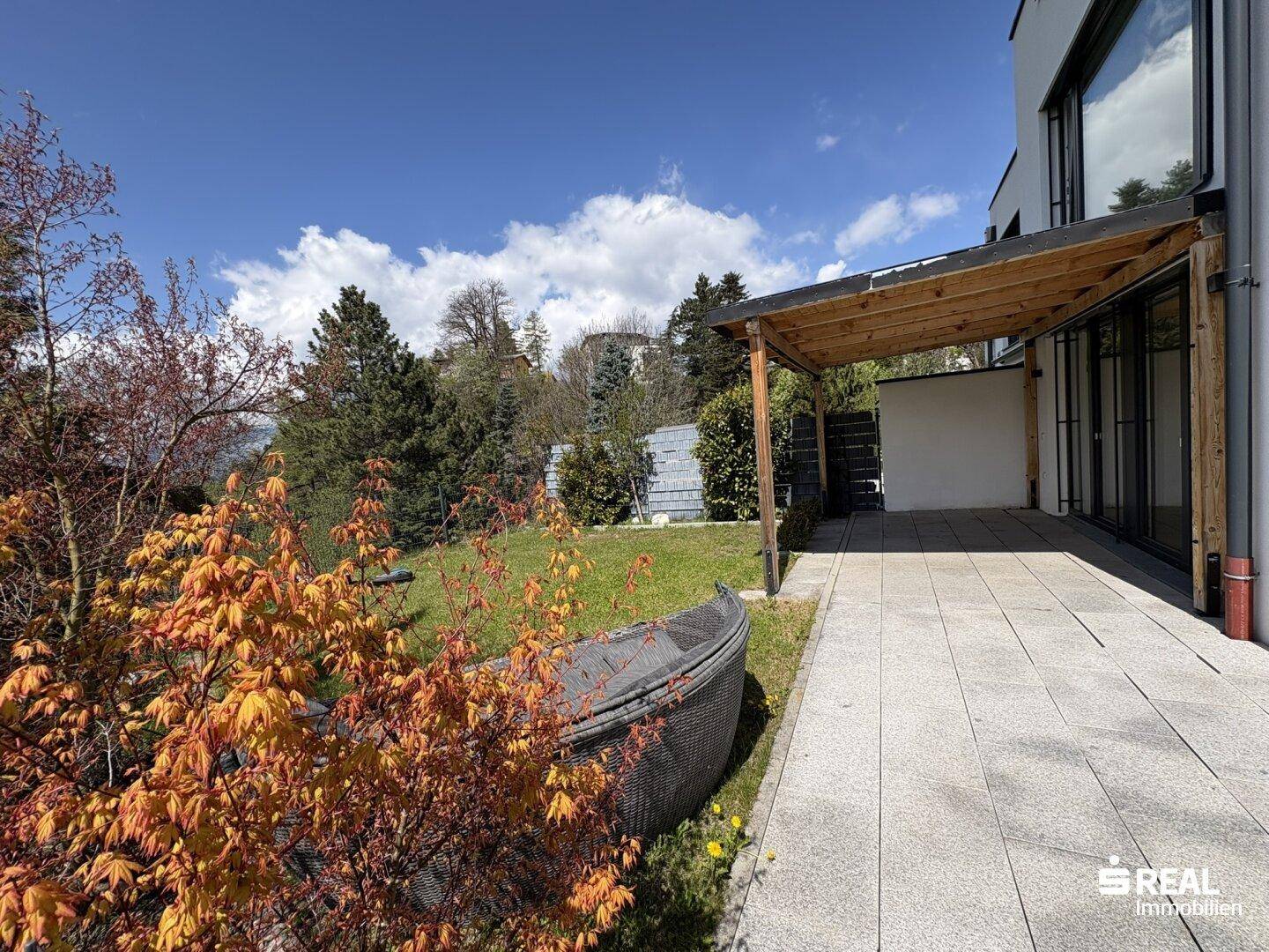 Sonniger Gartenbereich mit gepflasterter Terrasse, Pergola und gemütlicher Lounge-Ecke.
