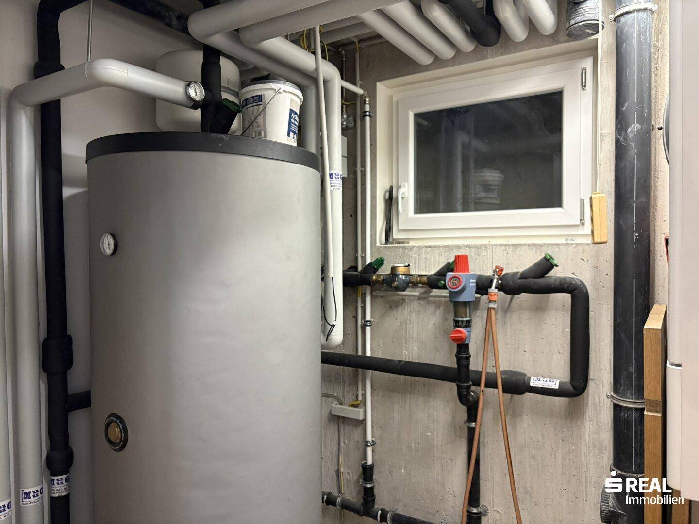 Technikraum mit Warmwasserspeicher, Heizungsrohren und kleinem Fenster.
