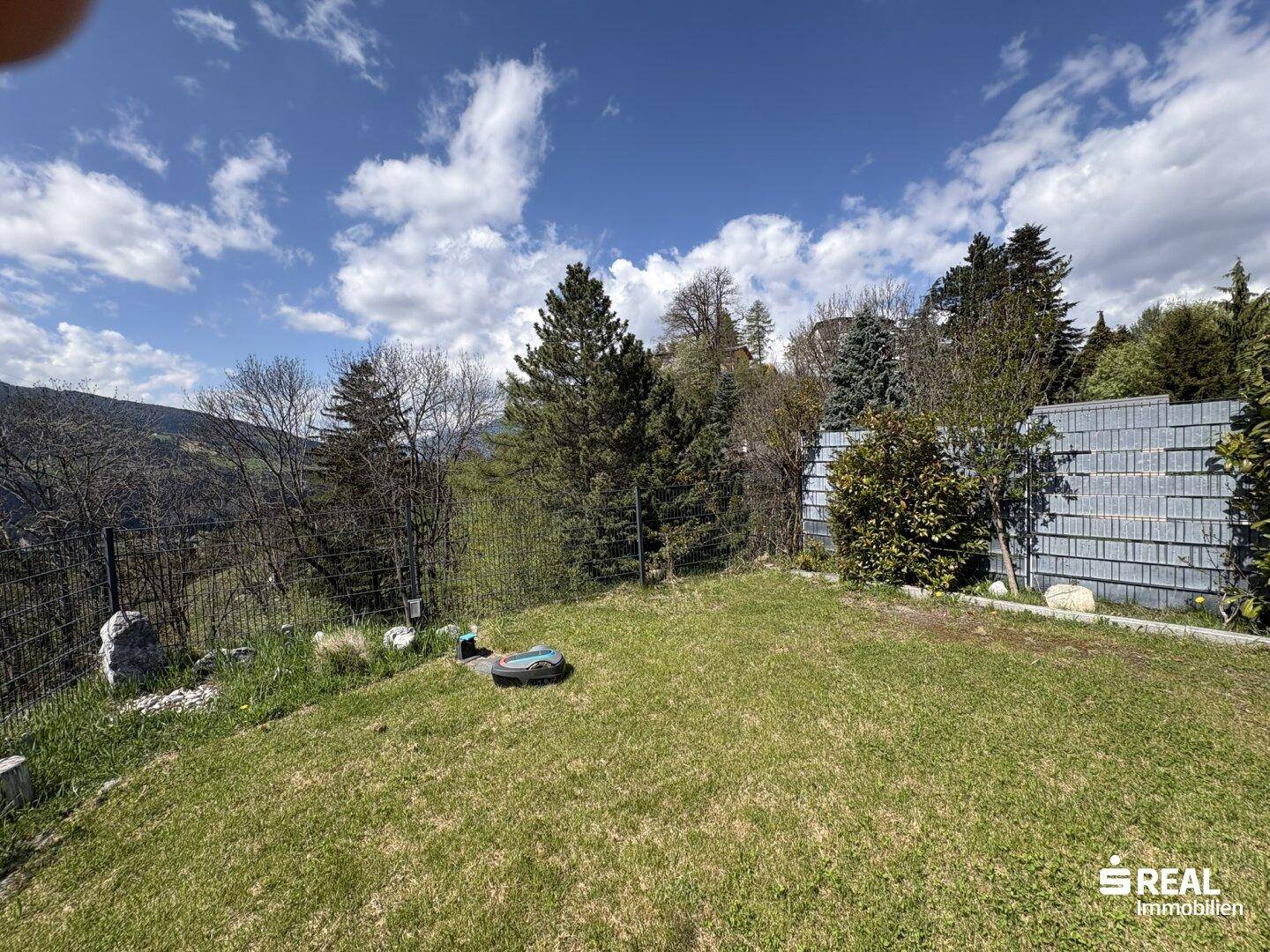 Gepflegter Garten mit Rasenfläche und umzäuntem Bereich, umgeben von Bäumen.