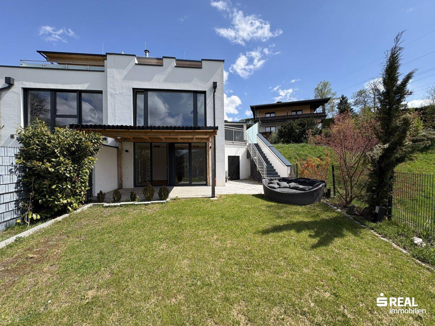 Moderner Neubau mit großem Garten, überdachter Terrasse und Außenwhirlpool unter blauem Himmel.