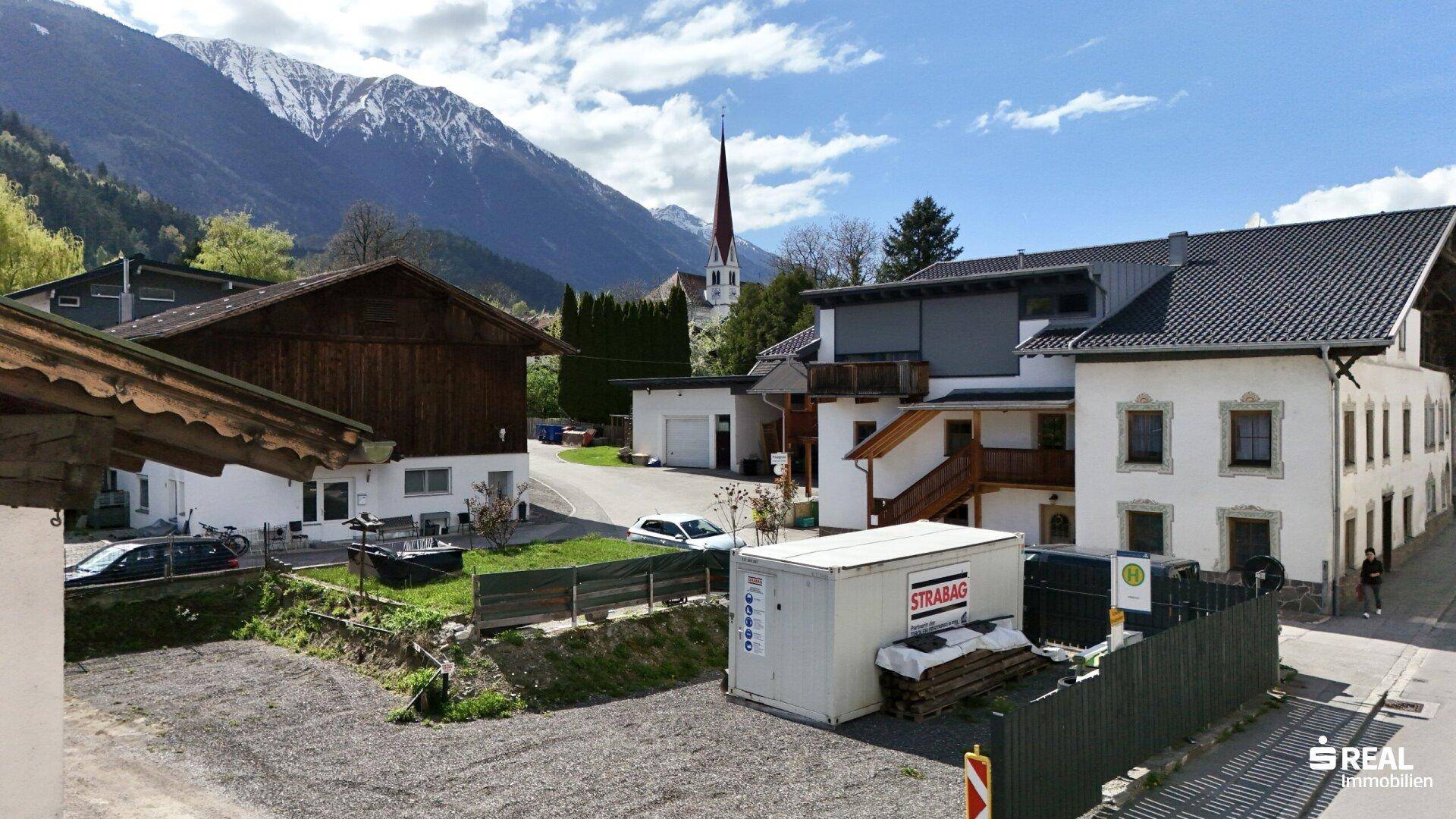 Blick auf traditionelle Gebäude in einer ländlichen Umgebung mit majestätischen Bergen im Hintergrund.