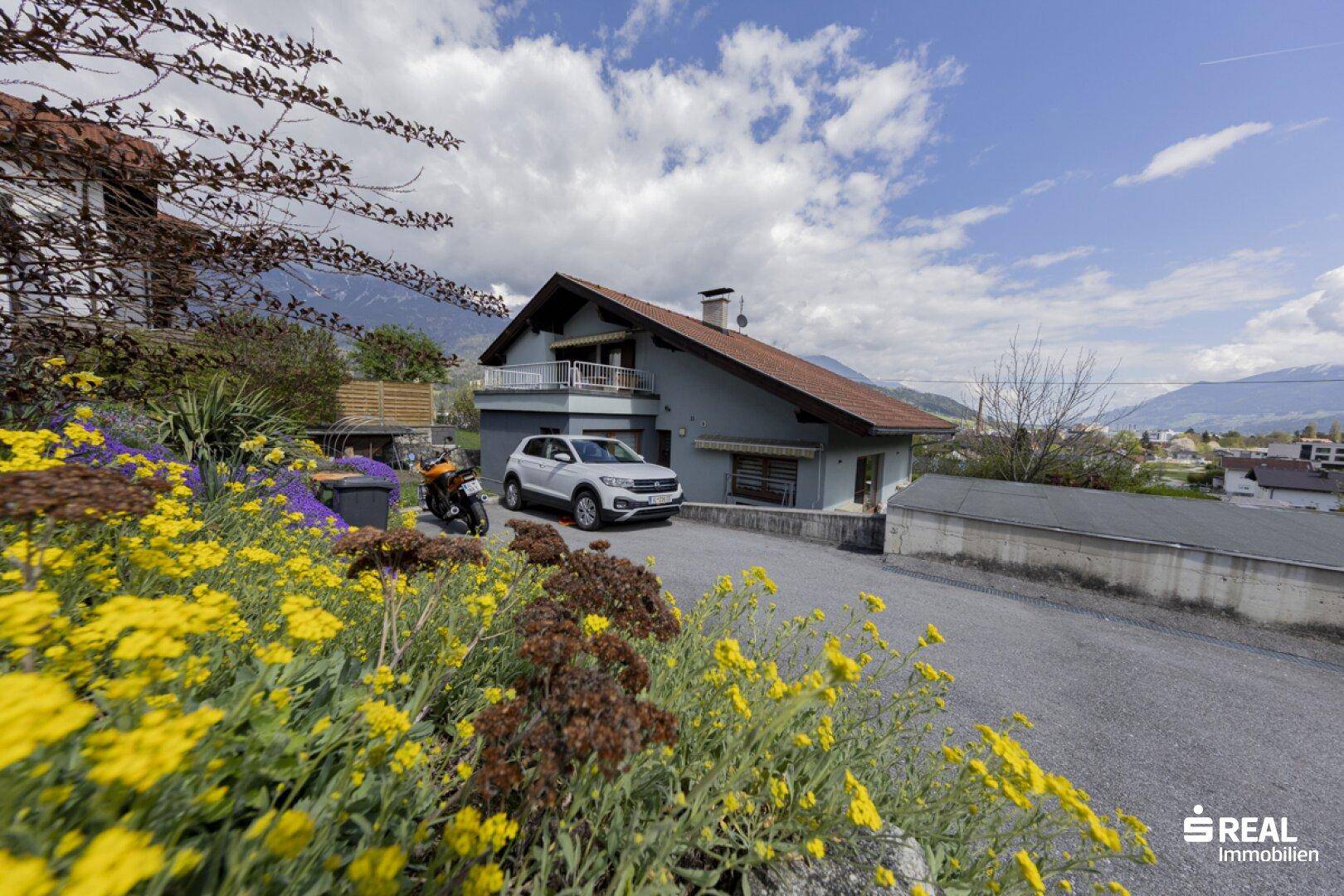 Hausfassade mit Einfahrt, Stellplatz und blühenden Pflanzen im Vorgarten mit Bergblick.