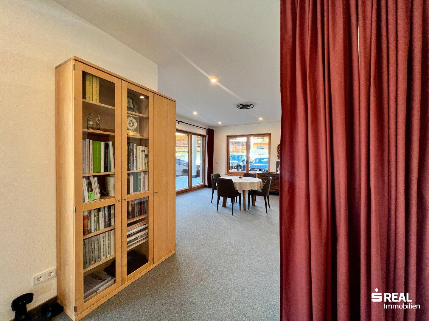 Heller Wohnbereich mit Bücherregal und Blick auf den Essbereich mit großen Fenstern.