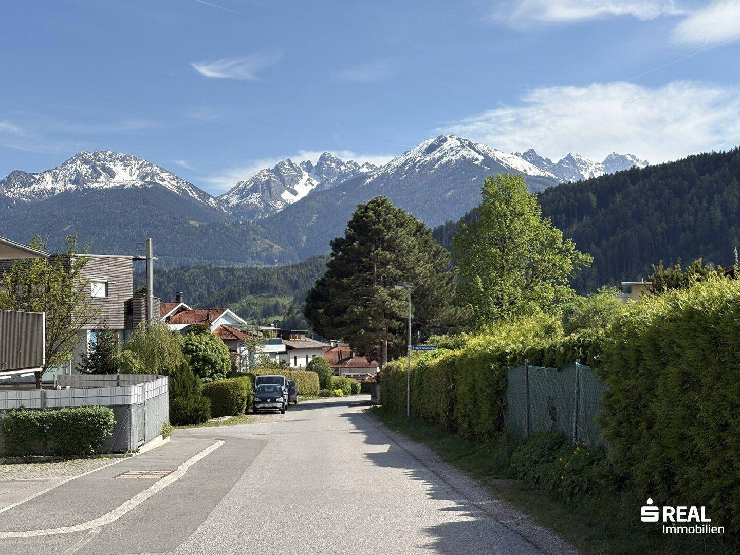 Ruhige Wohnstraße mit grünen Hecken und Häusern, umrahmt von einer beeindruckenden Berglandschaft.