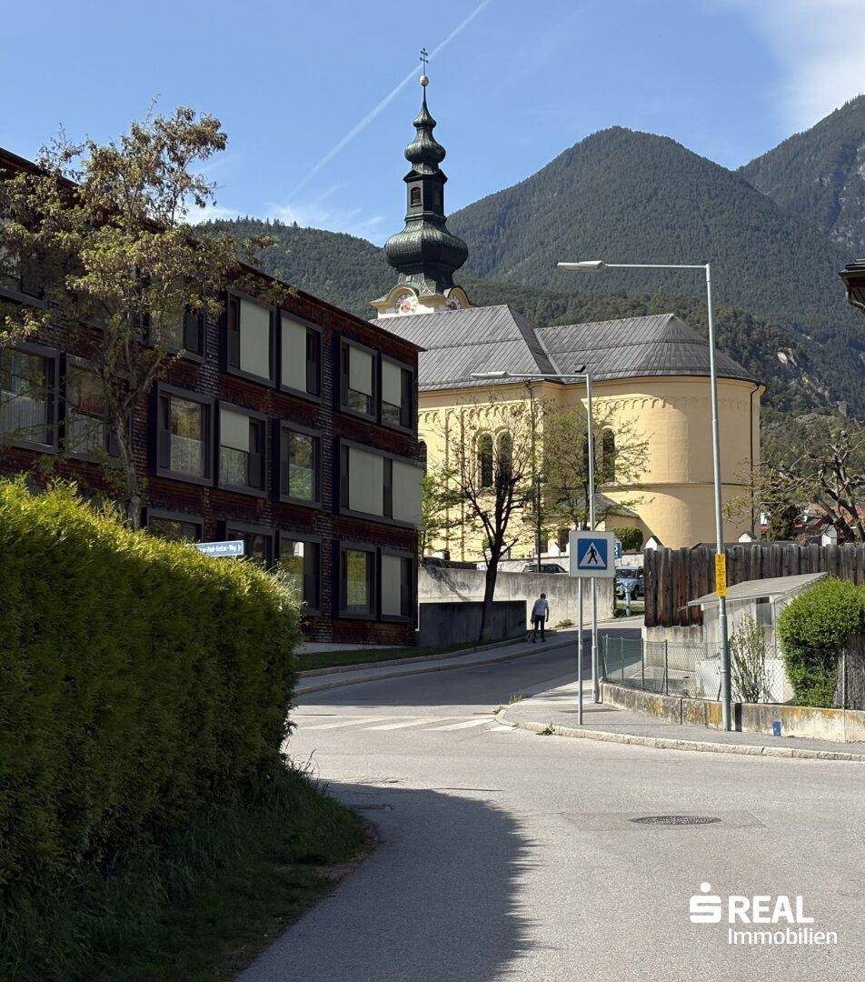 Straßenansicht mit einer historischen Kirche und einem modernen Gebäude vor einer Bergkulisse.