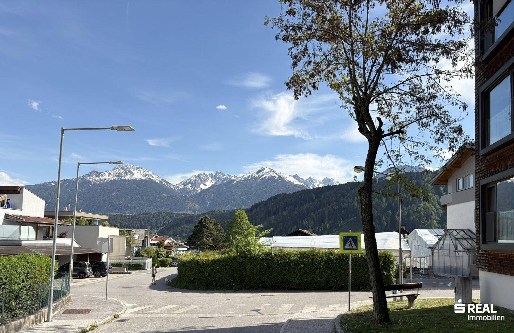 Straßenkreuzung mit einem Baum und Häusern, im Hintergrund erheben sich majestätische Berge.