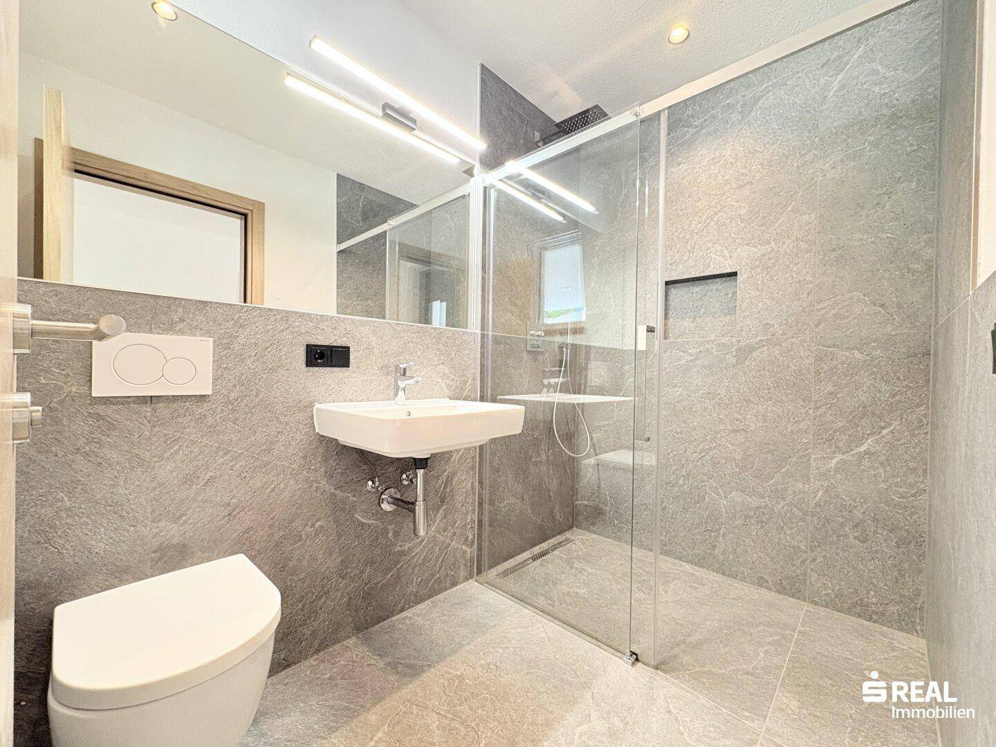 Modernes Badezimmer mit grauen Fliesen, wandhängendem WC, Einzelwaschbecken und begehbarer Dusche.