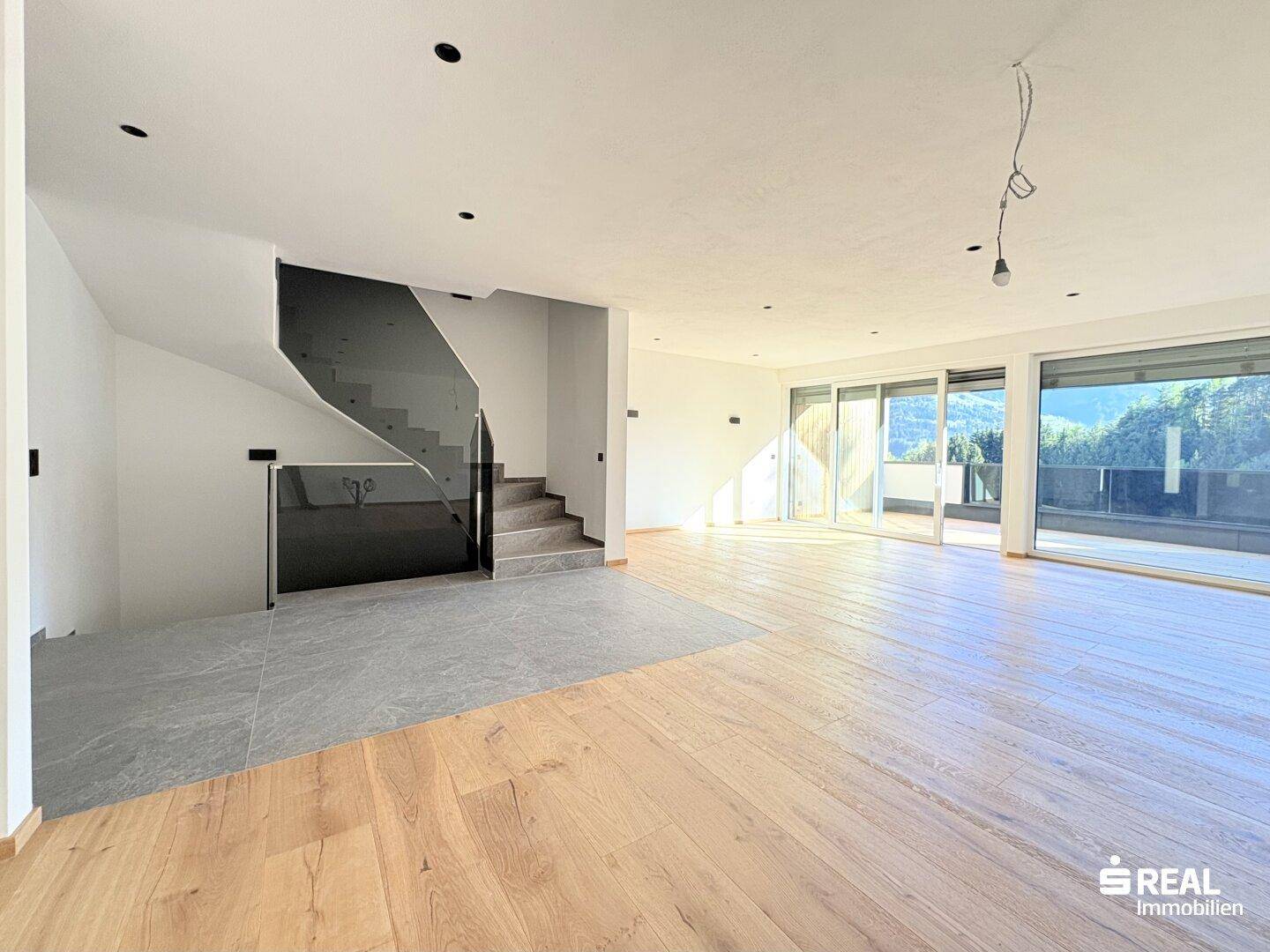 Offener Wohnbereich mit Holzboden, großen Fenstern und einer modernen Treppe mit Glasgeländer.