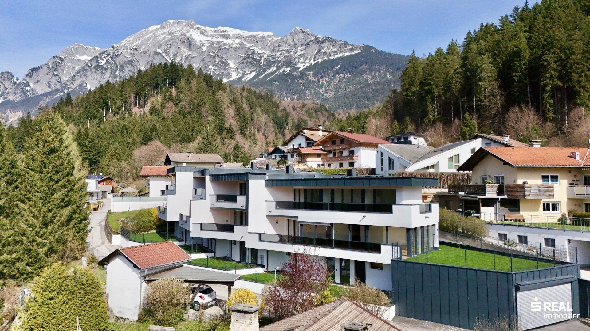 Moderne Wohnanlage in Hanglage mit weißer Fassade und großen Fensterfronten vor Bergkulisse.