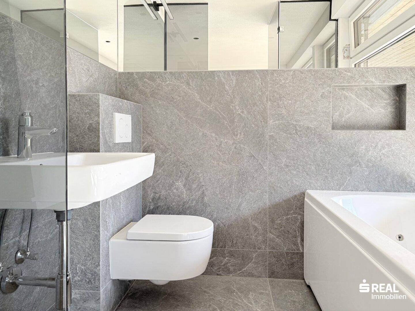 Modernes Badezimmer mit grauen Fliesen, wandhängendem WC und integrierter Badewanne.
