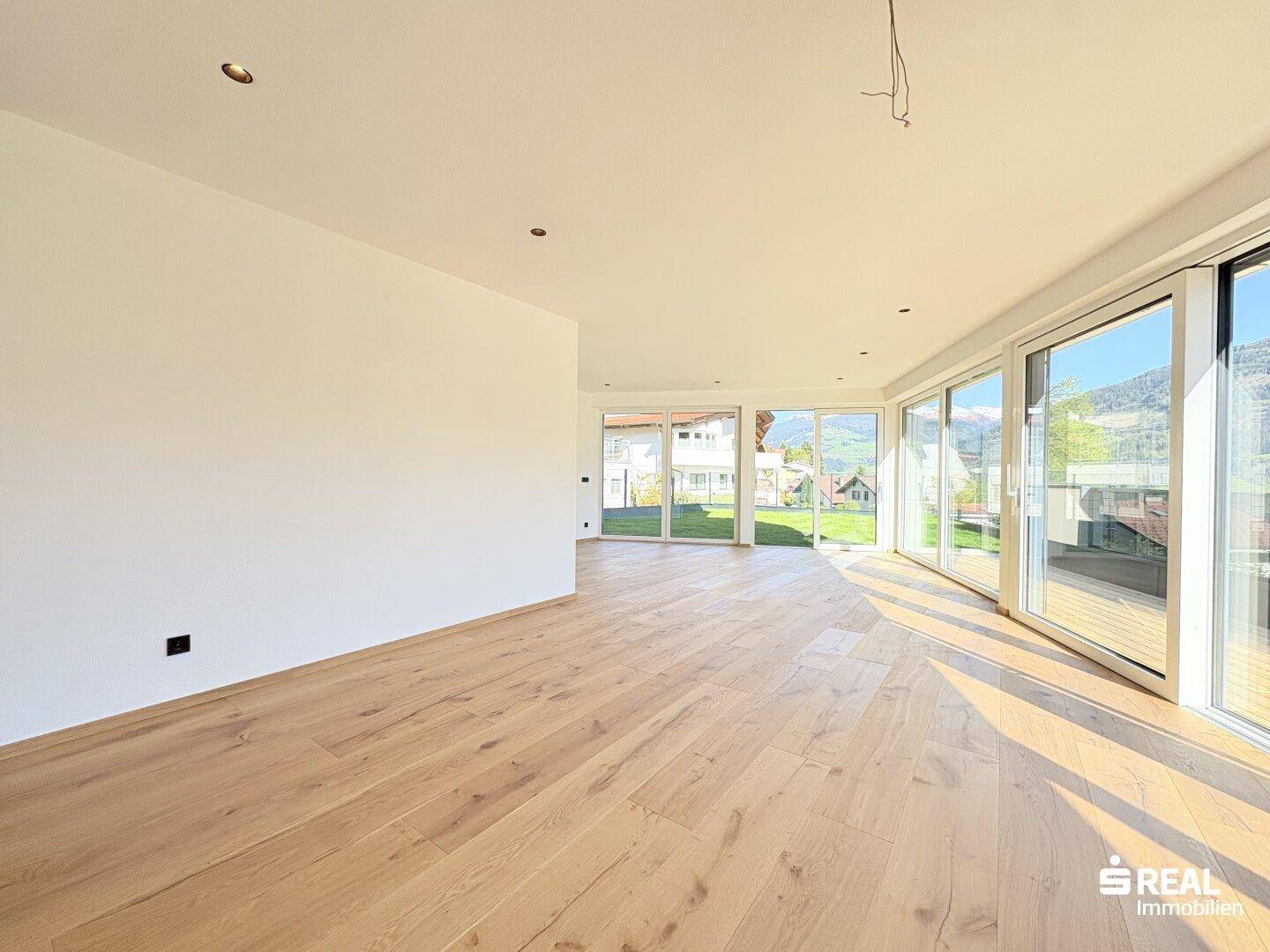Heller, leerer Wohnraum mit großen Fensterfronten und hellem Holzparkettboden.
