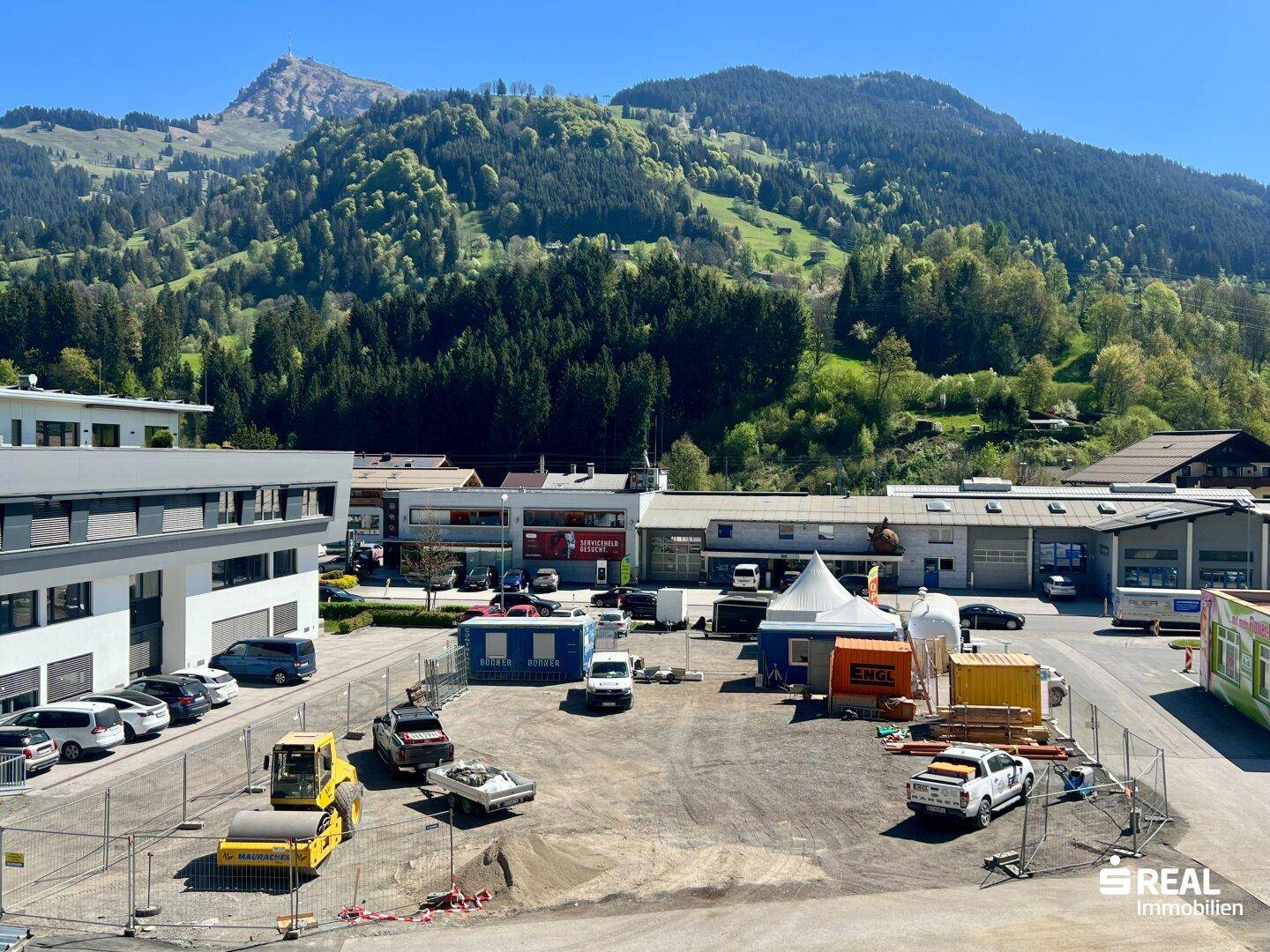 Nahaufnahme eines Grundstücks mit Baucontainern und Fahrzeugen, umgeben von Bergen und Bäumen.