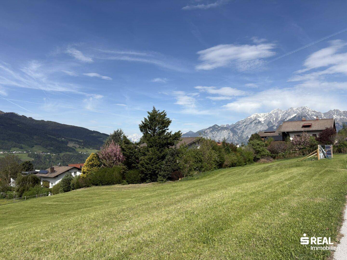 Weitläufige grüne Wiese mit Blick auf die umliegenden Häuser und die majestätische Bergkulisse.