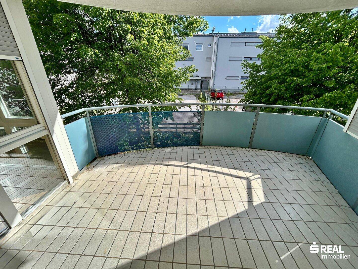 Sonniger Balkon mit Fliesenboden und Blick auf Bäume und umliegende Gebäude.