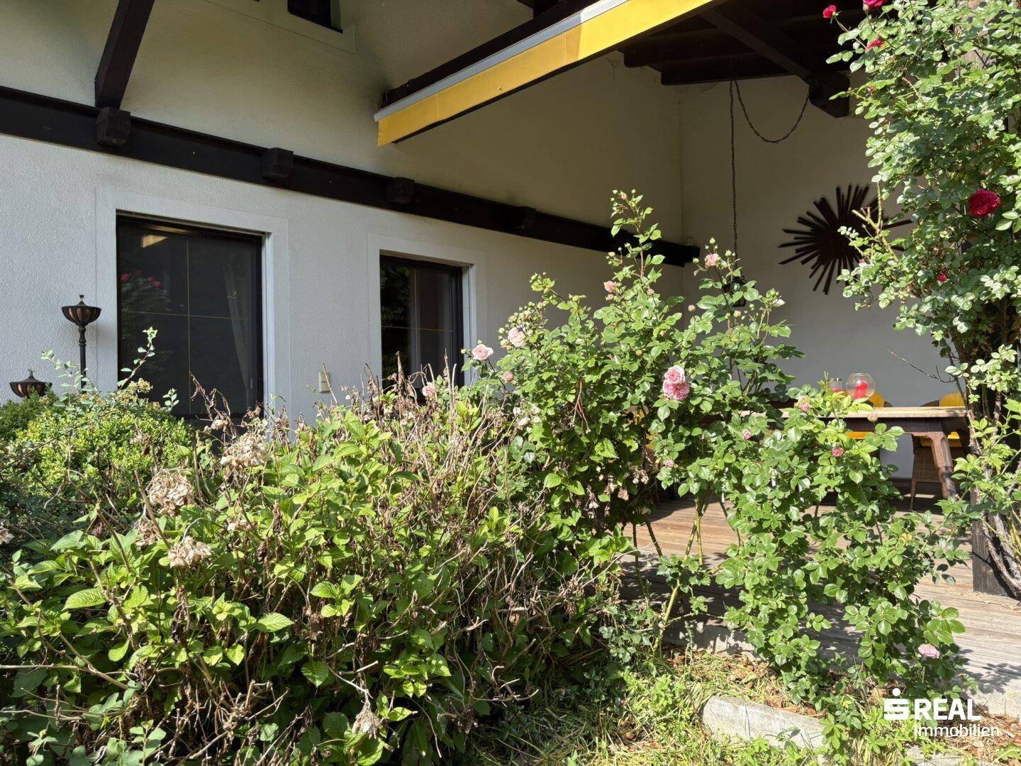 Terrasse mit Markise und blühenden Pflanzen, umgeben von viel Grün.
