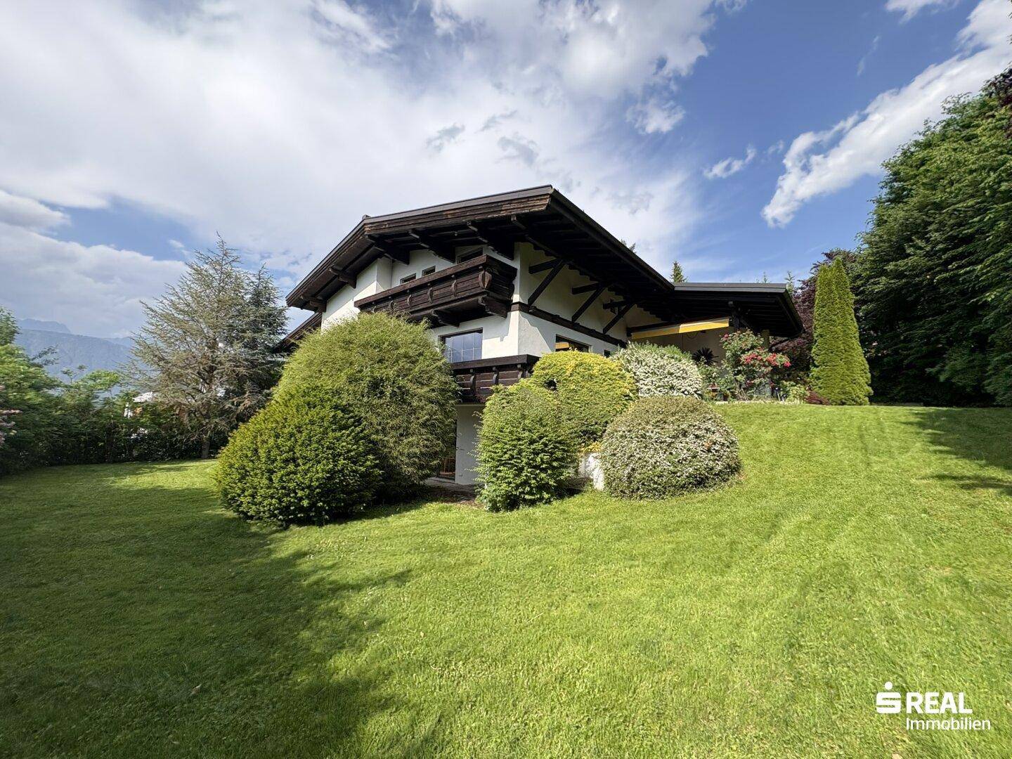 Charmantes Haus im alpenländischen Stil mit weitläufigem Rasen und üppiger Bepflanzung unter blauem Himmel.