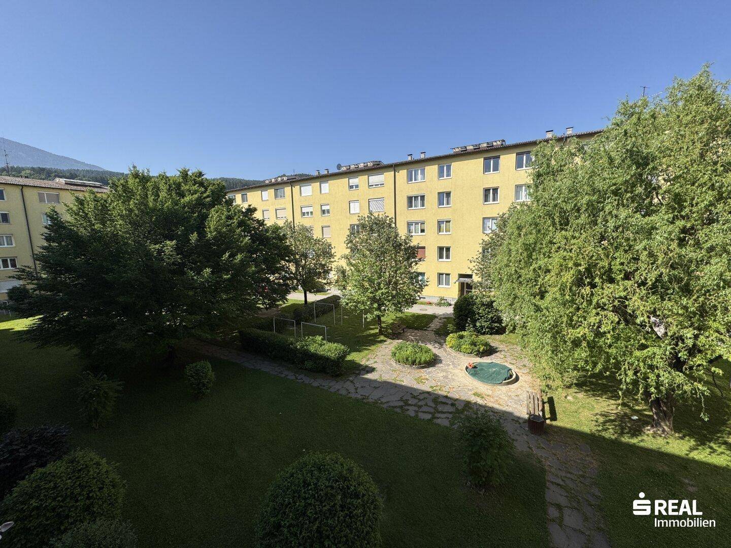 Sonniger Blick vom Balkon auf den gepflegten Innenhof mit Bäumen, Sträuchern und einem kleinen Wasserbecken.