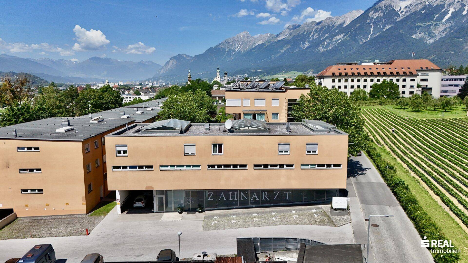 Gebäudeansicht der Zahnarztpraxis mit angrenzenden Feldern und weitem Blick auf die Stadt und Berge.
