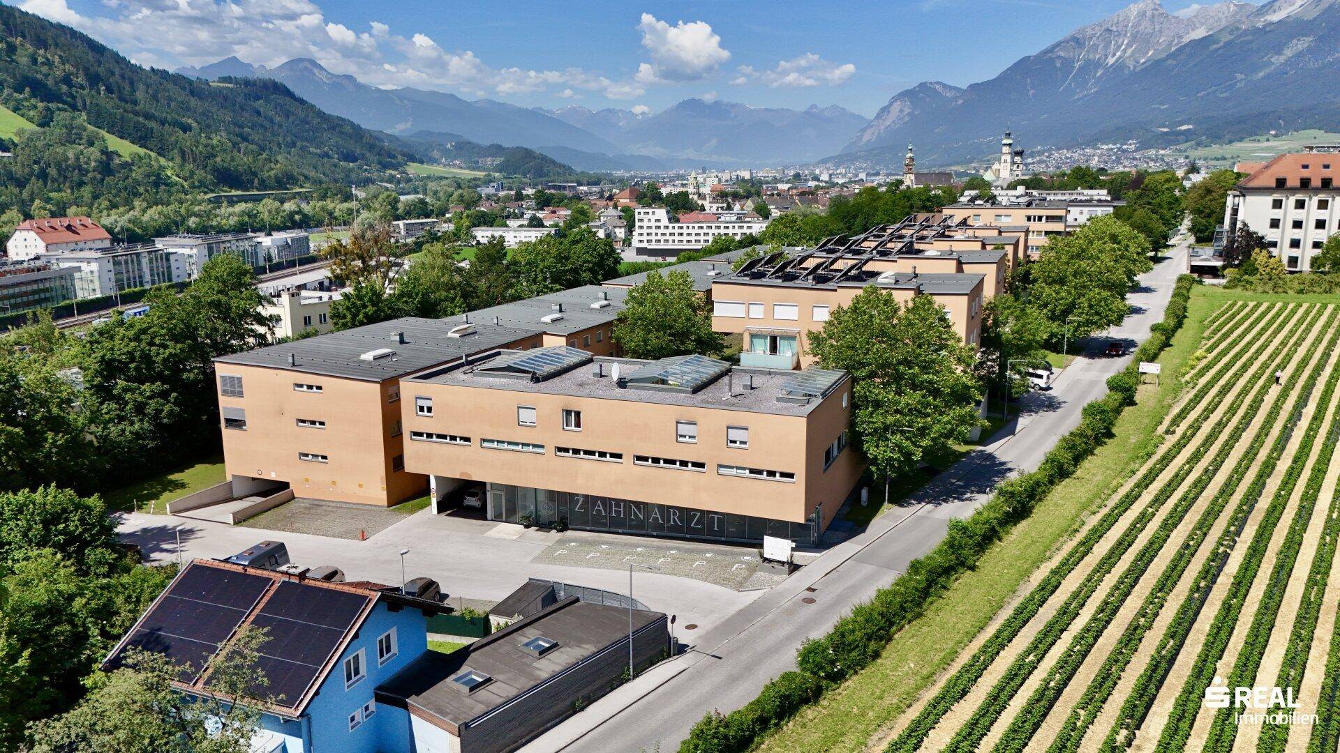 Luftaufnahme des Gebäudekomplexes mit Parkplätzen, angrenzenden Feldern und der Berglandschaft.