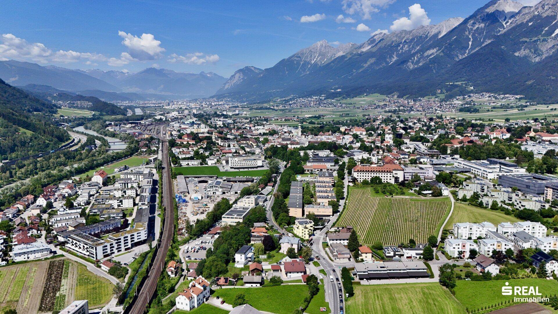 Weitläufige Luftaufnahme eines Tals mit Stadt, Fluss, Bahnlinie und majestätischer Bergkulisse.