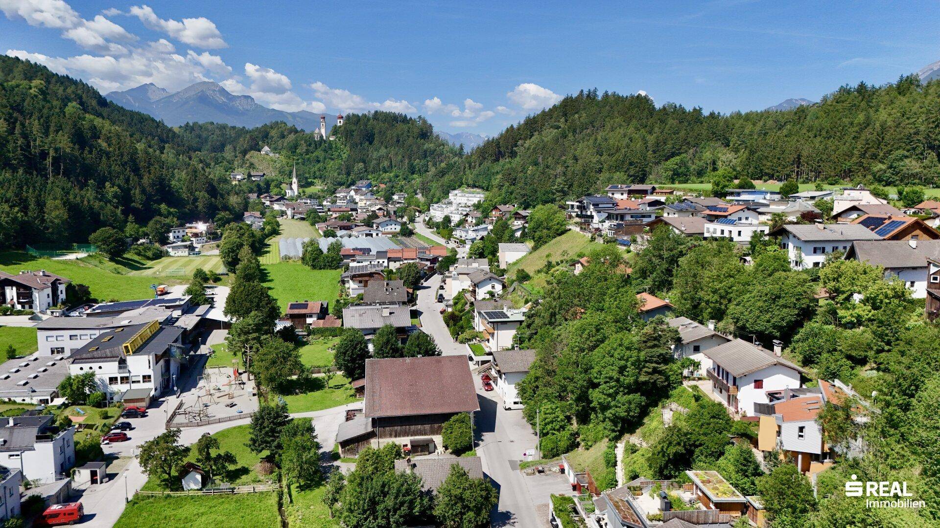 Weitläufige Luftaufnahme eines malerischen Dorfes eingebettet in eine grüne Hügellandschaft mit Bergen.
