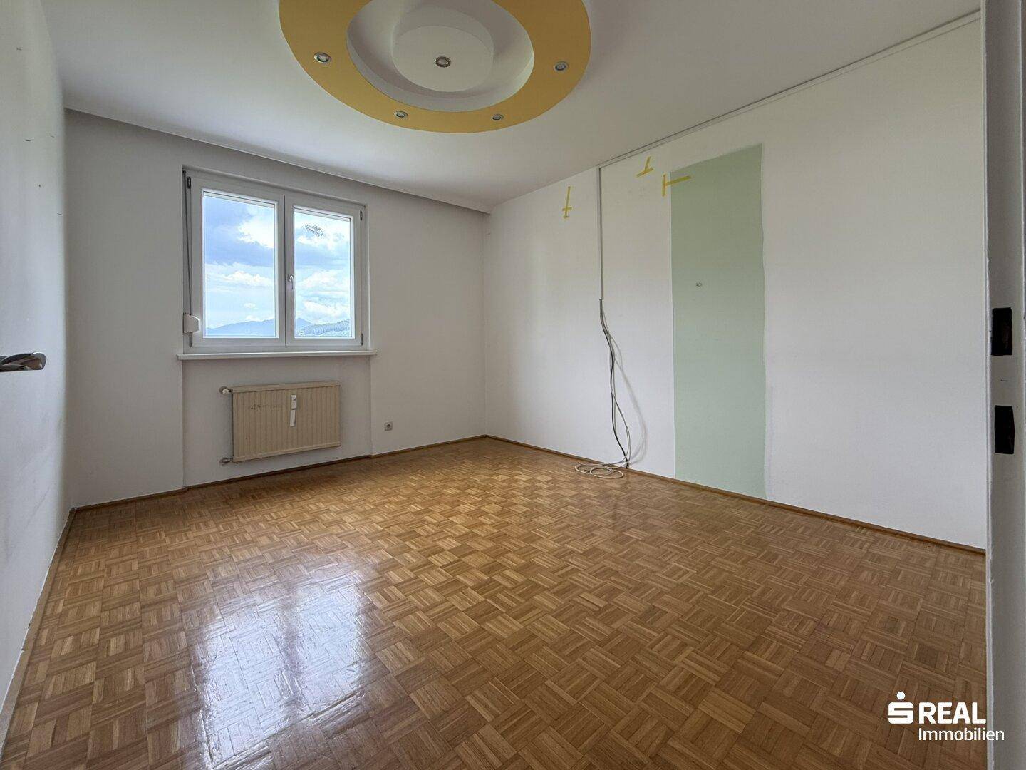 Leeres Zimmer mit Parkettboden, Fenster und einer dekorativen Deckenbeleuchtung in Gelb und Weiß.