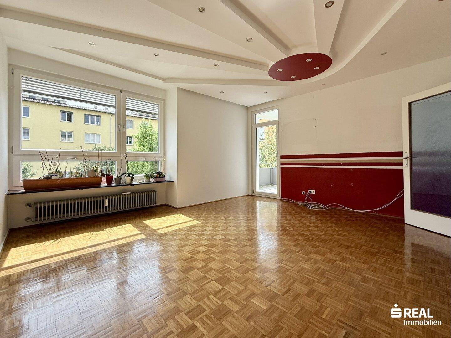 Geräumiges Wohnzimmer mit Parkettboden, großen Fenstern und Zugang zu einem kleinen Balkon.