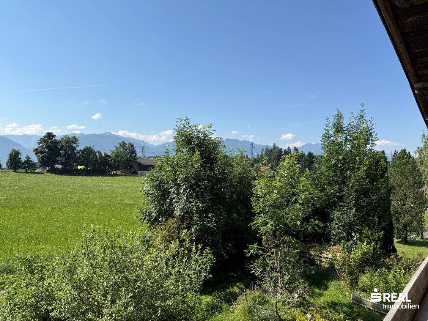 Panoramablick über grüne Bäume und Wiesen auf die umliegende Berglandschaft an einem sonnigen Tag.